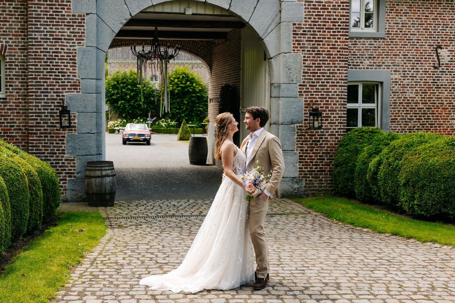 Bride and groom share a romantic moment on cobblestone courtyard outside a grand brick estate with arched entrance.
