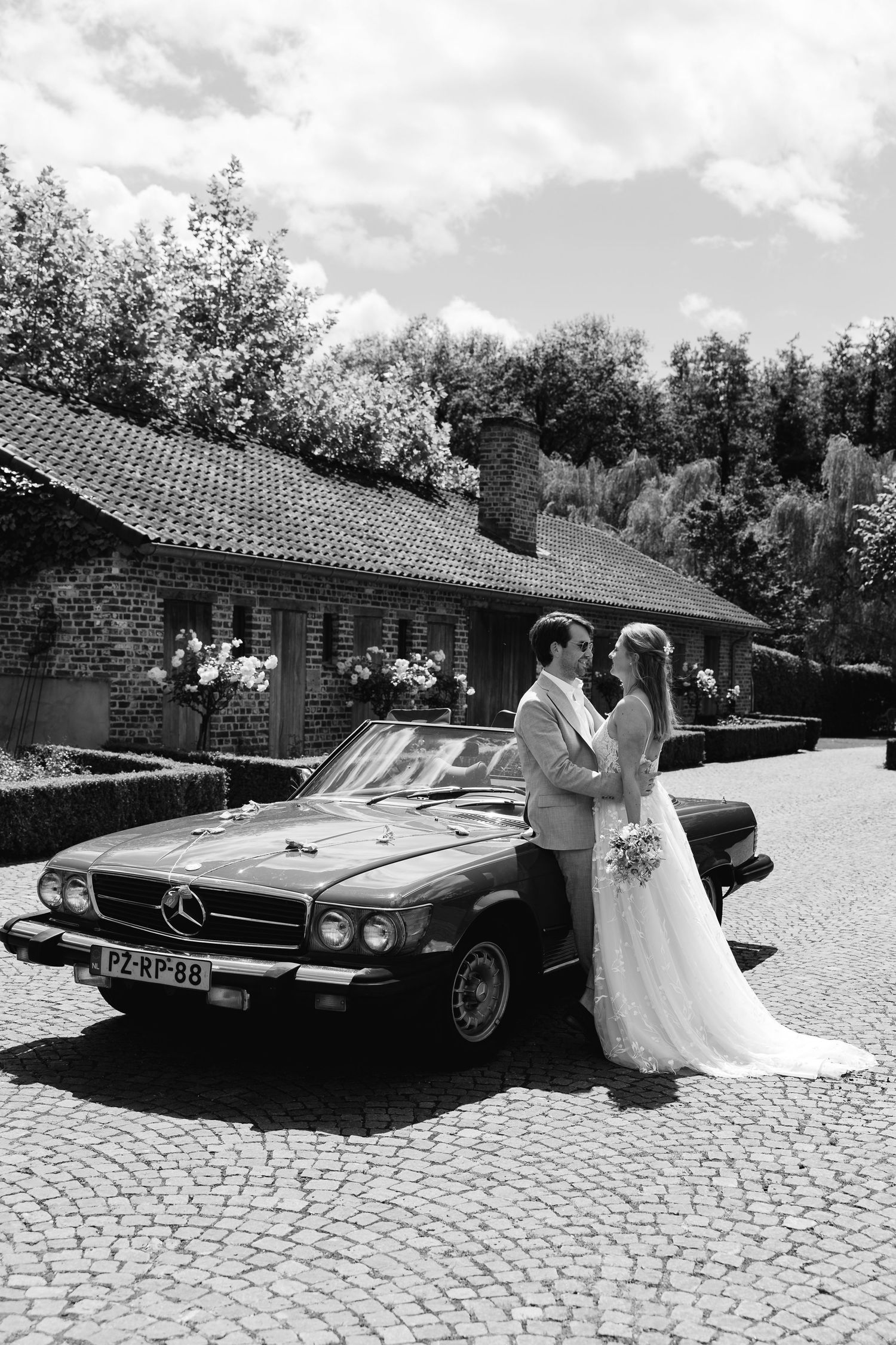 Bride and groom pose beside a classic vintage Mercedes convertible on cobblestone driveway at wedding venue.