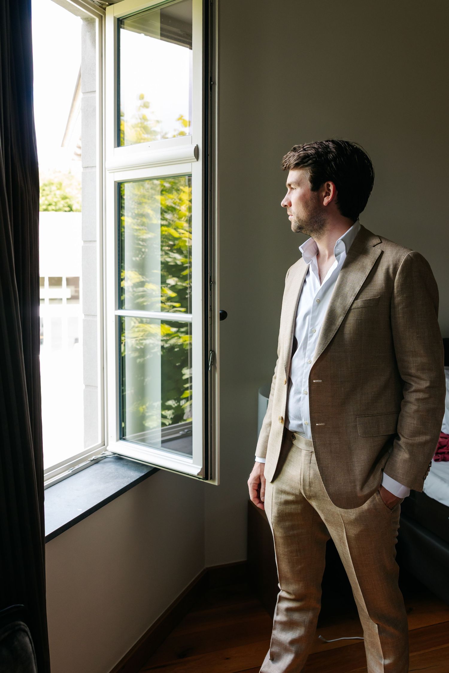 A well-dressed man in a tan suit gazes thoughtfully out an open window in a dimly lit room.