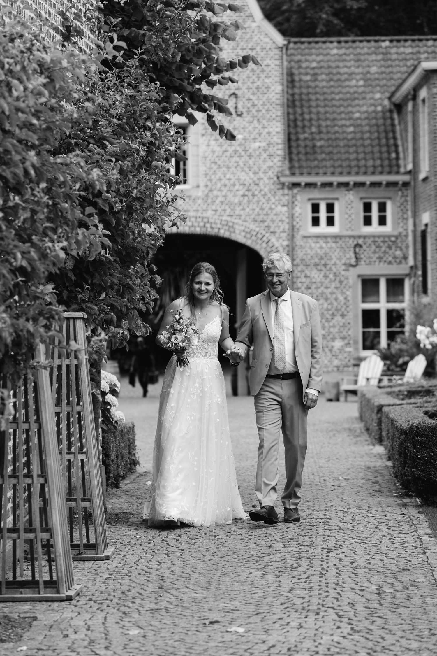 Black and white photo of bride walking with her father along a cobblestone path at a historic venue.