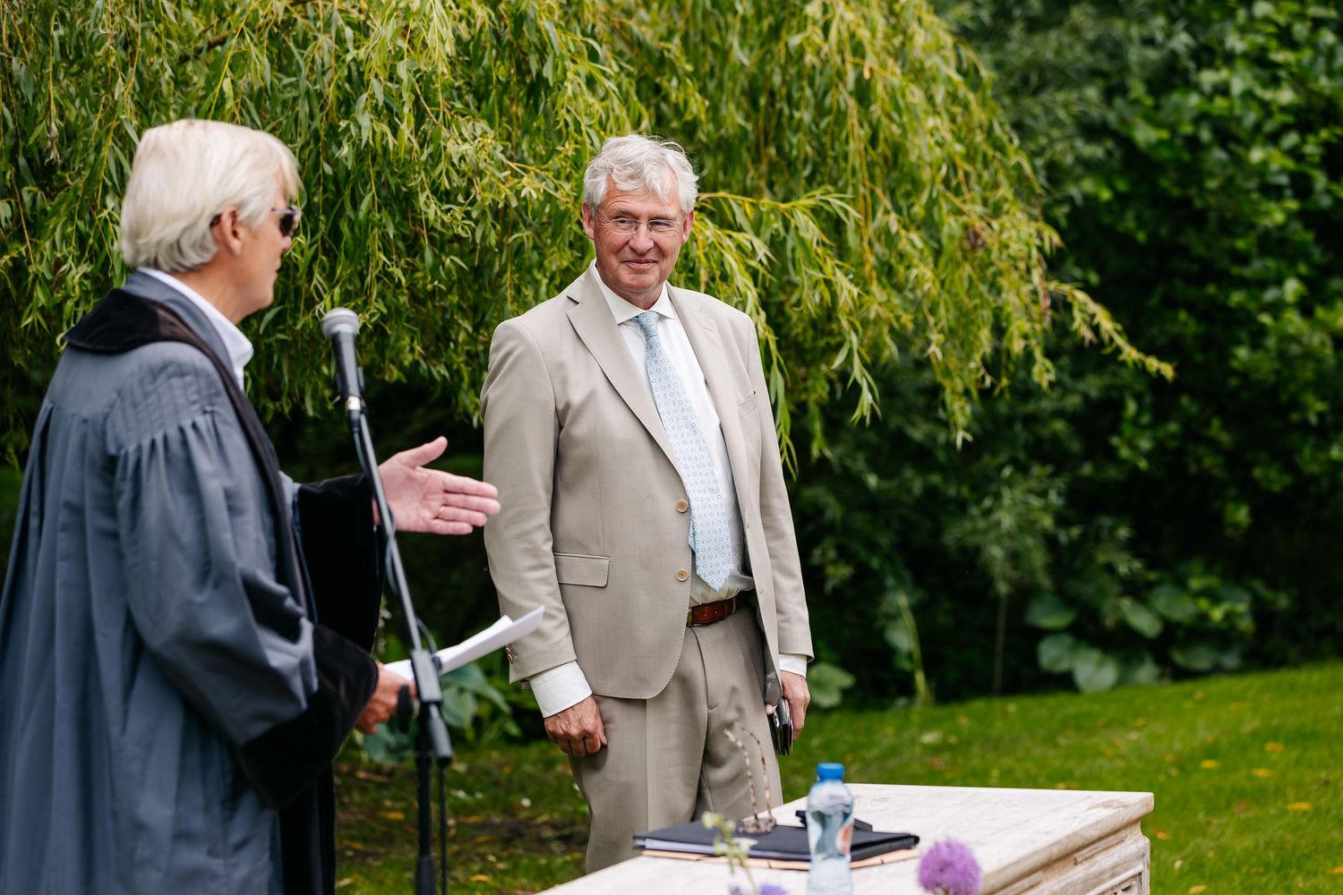 Two men in suits converse outdoors at a garden ceremony near a microphone stand and decorated table.