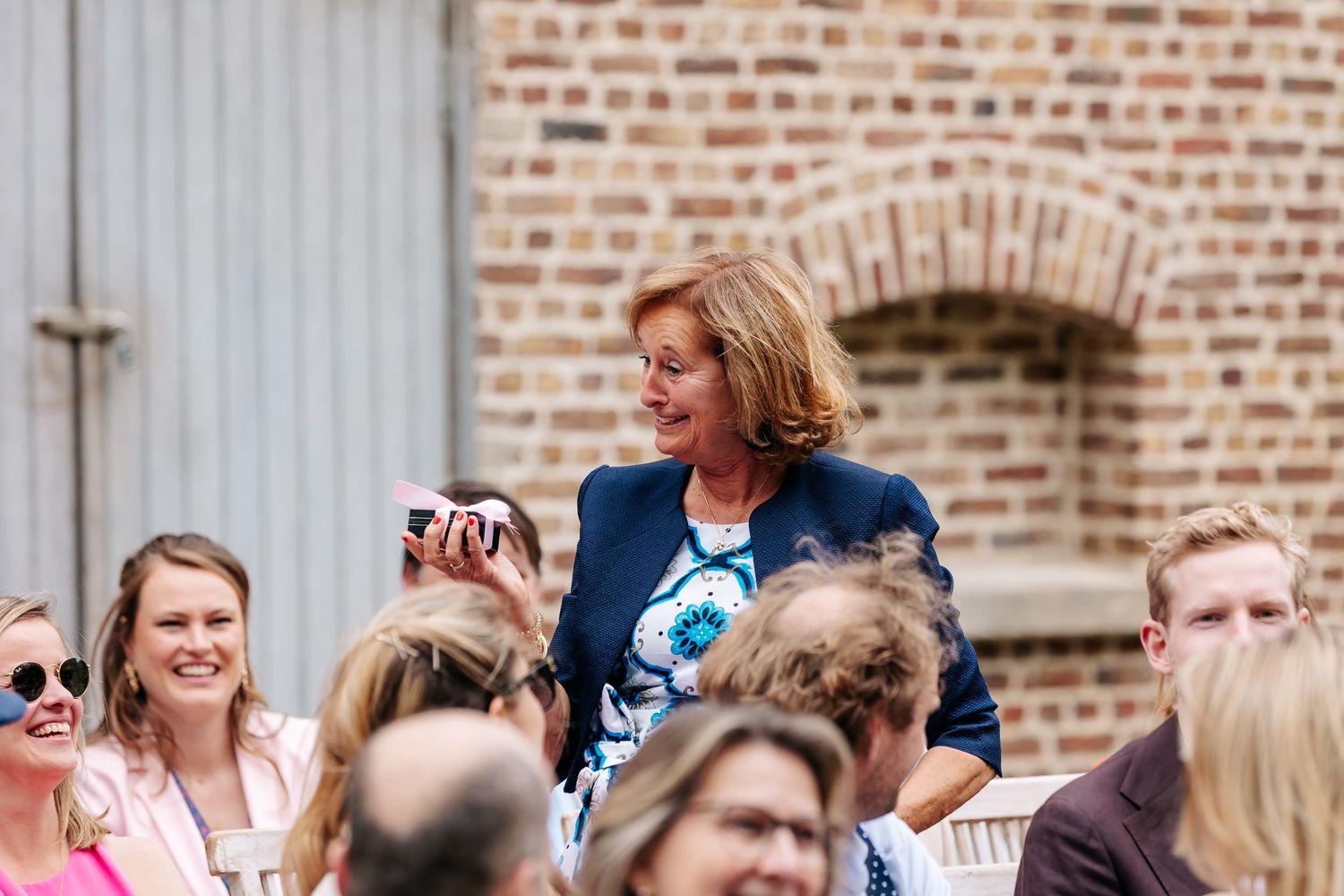 Elegantly dressed woman in navy blazer smiling among seated wedding ceremony guests.
