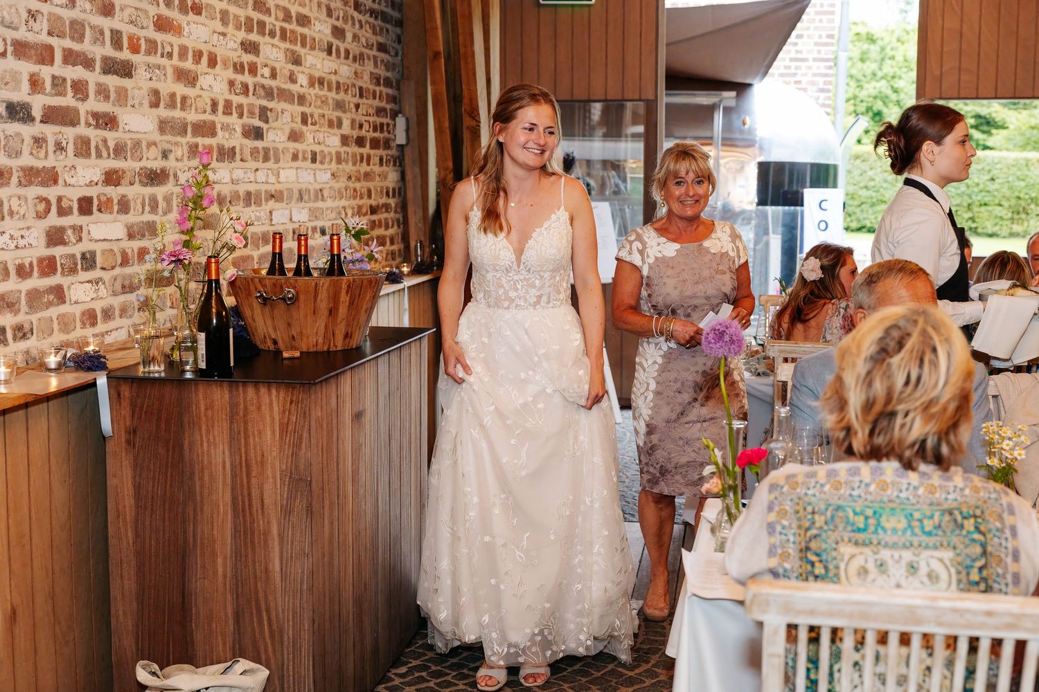 Smiling bride in white lace wedding gown entering reception venue accompanied by guests and staff.
