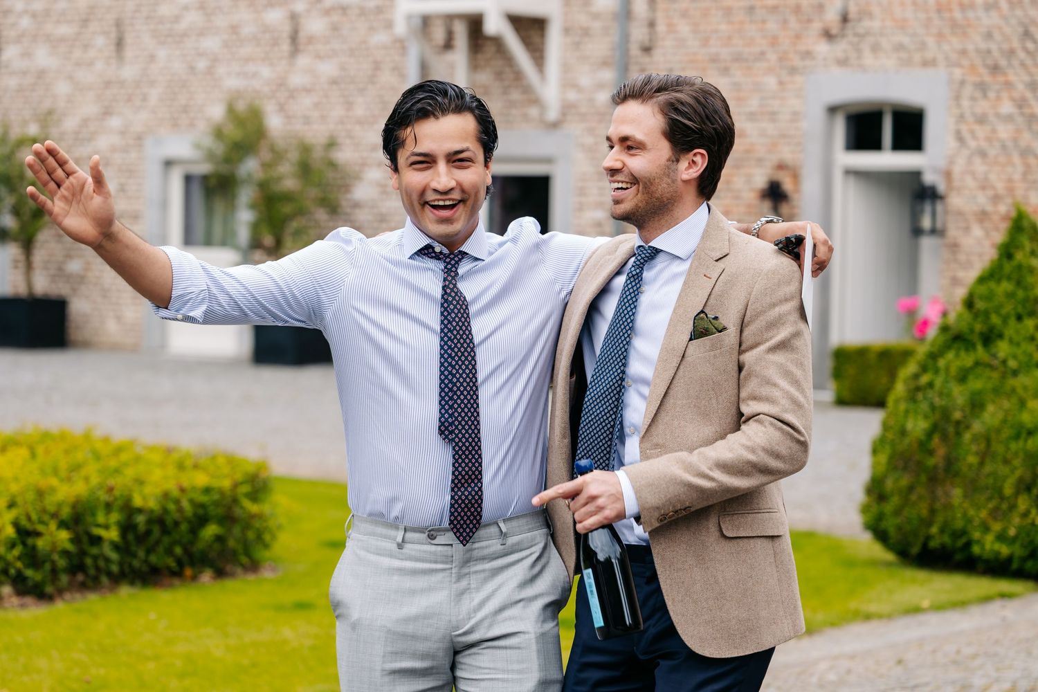 Two well-dressed men in suits and ties laughing and walking together outdoors at a wedding venue.