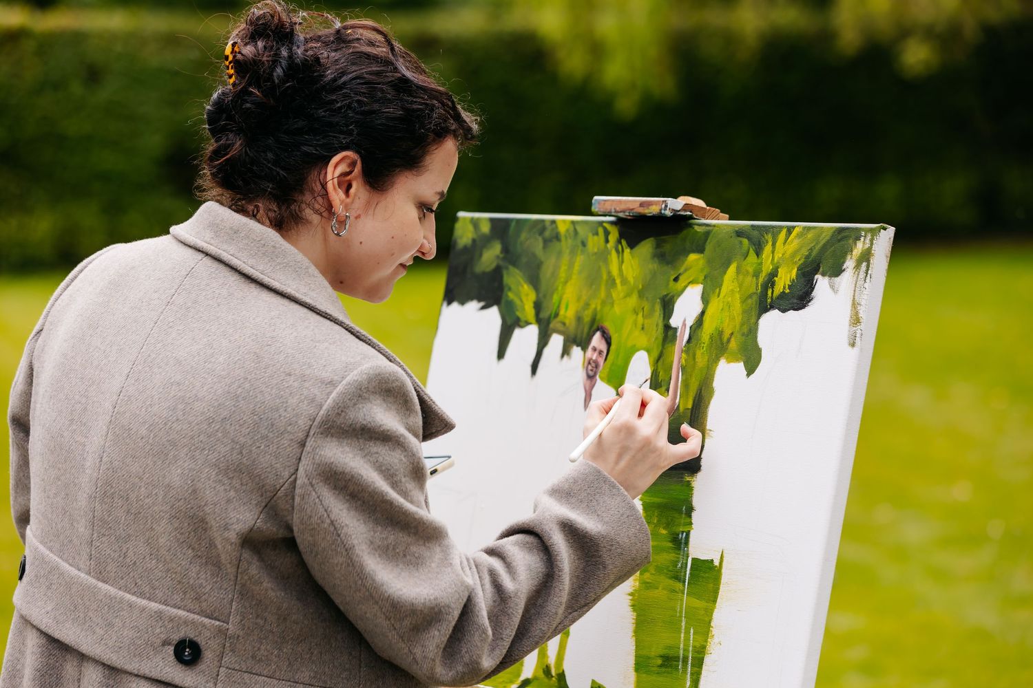 Woman in grey coat painting on canvas outdoors on an easel in a green garden.