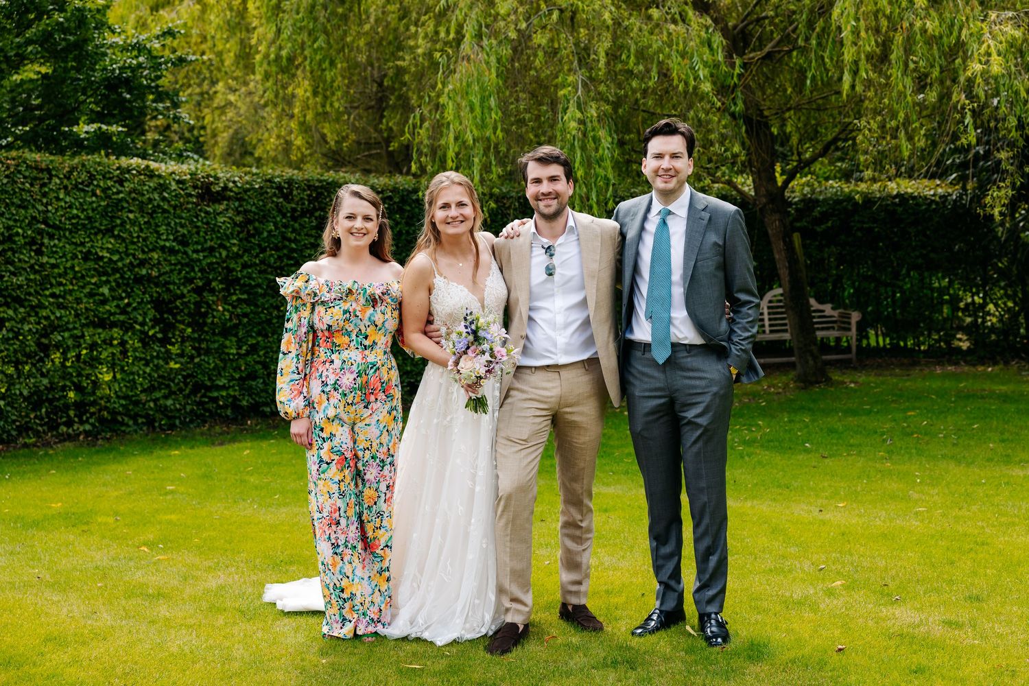 Bride and groom posing with two friends on green lawn, bride holding bouquet, guests in colorful summer attire.