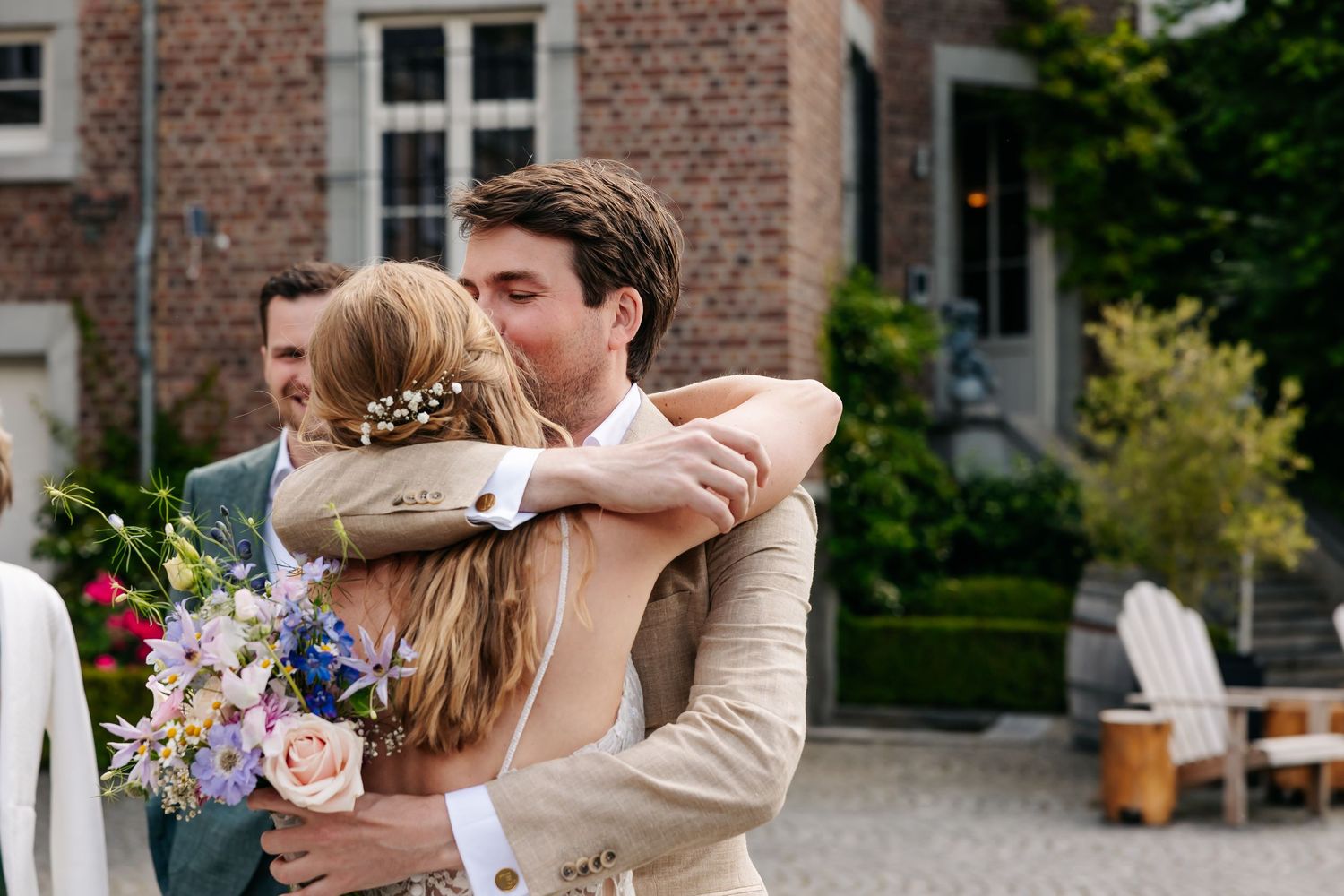 Groom in beige suit embracing bride holding colorful bouquet at outdoor wedding.