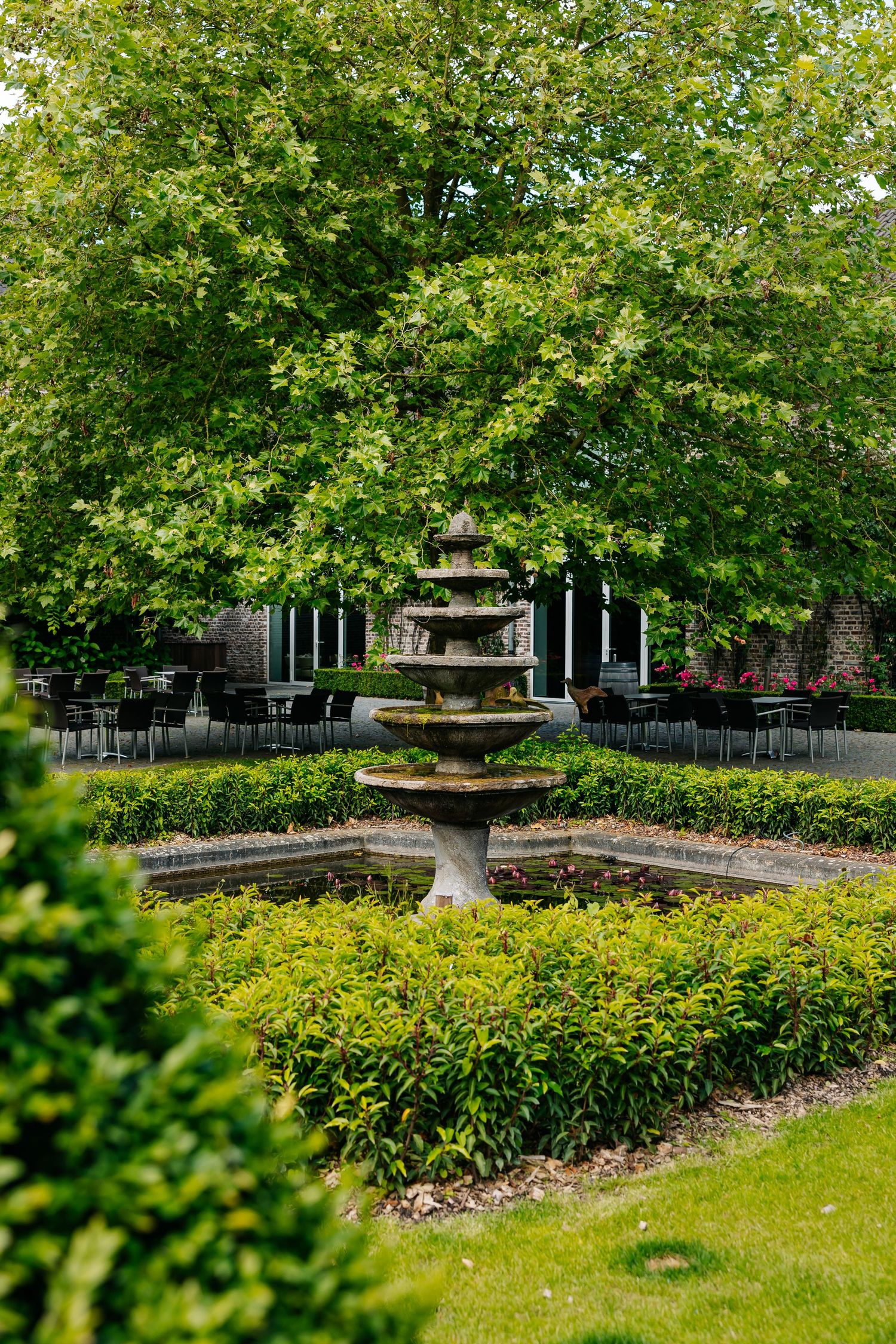 A tiered stone fountain surrounded by manicured hedges and lush green trees in an elegant garden setting.
