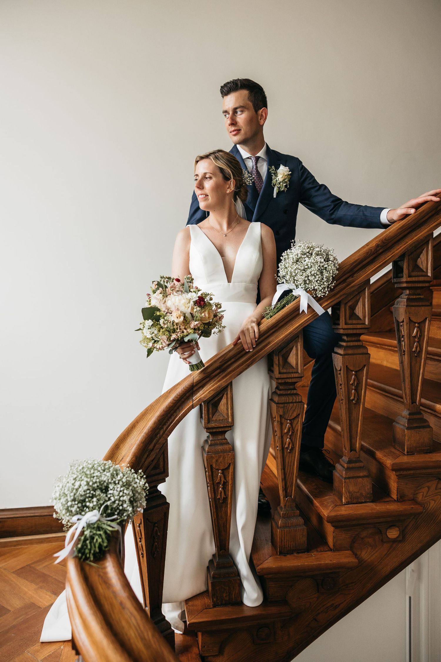 Elegant bride and groom posing on a grand wooden staircase adorned with baby's breath bouquets.