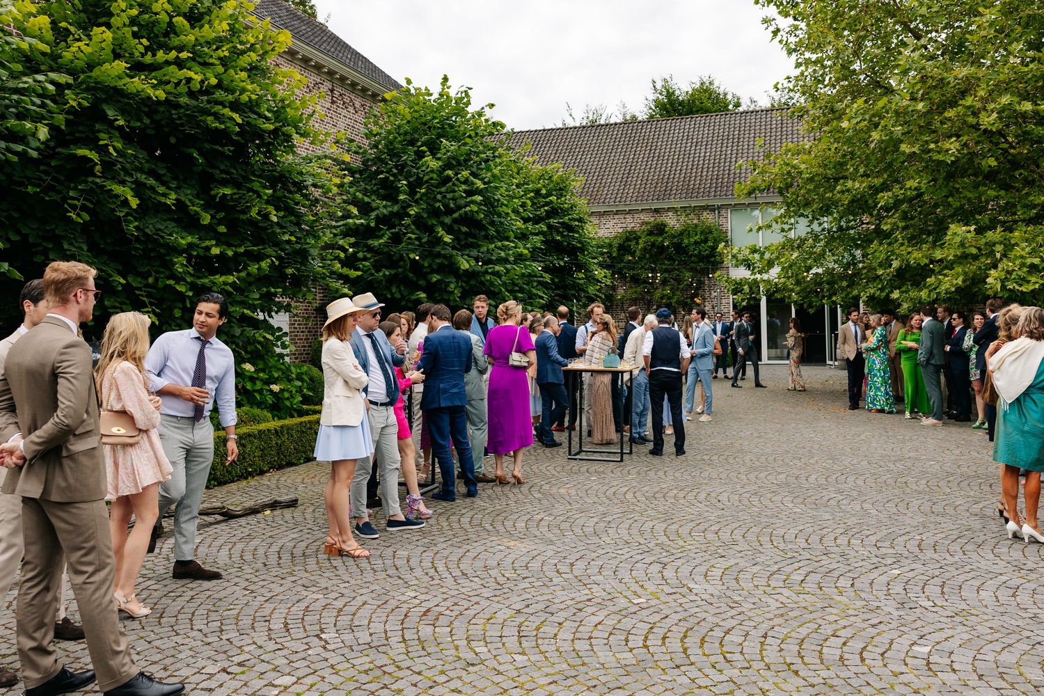 Wedding guests mingling on cobblestone courtyard surrounded by lush greenery at elegant venue.