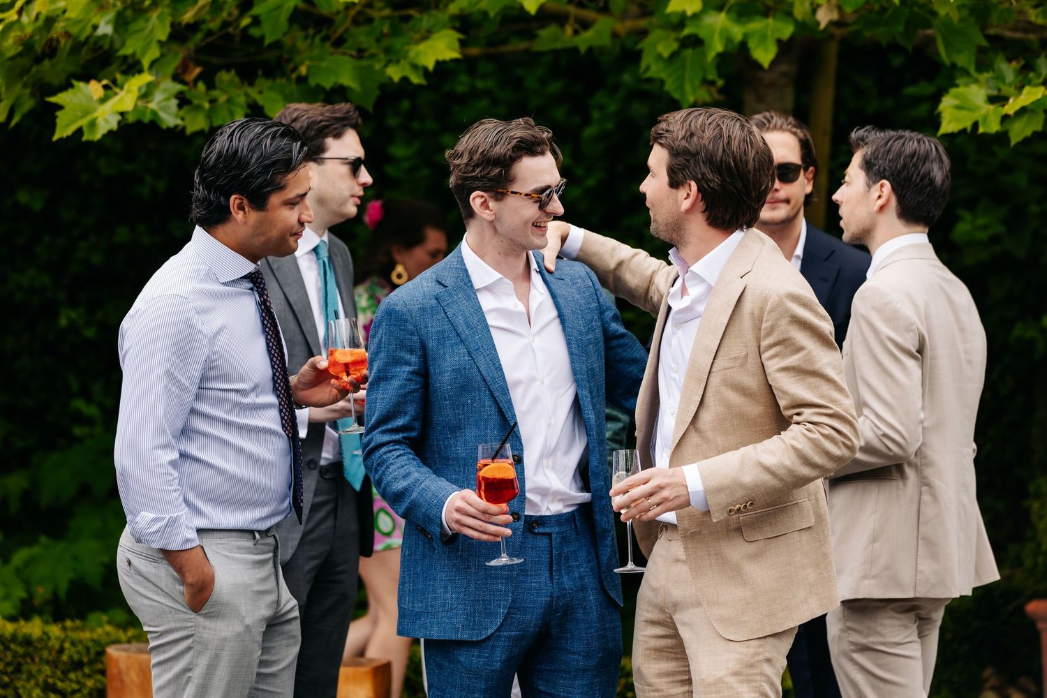 Group of men in elegant suits socializing and holding drinks at an outdoor wedding.