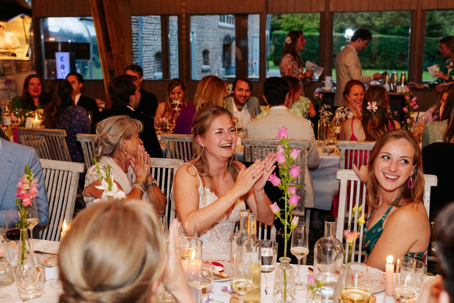 Bride and wedding guests laugh and clap at candlelit reception dinner in elegant venue.