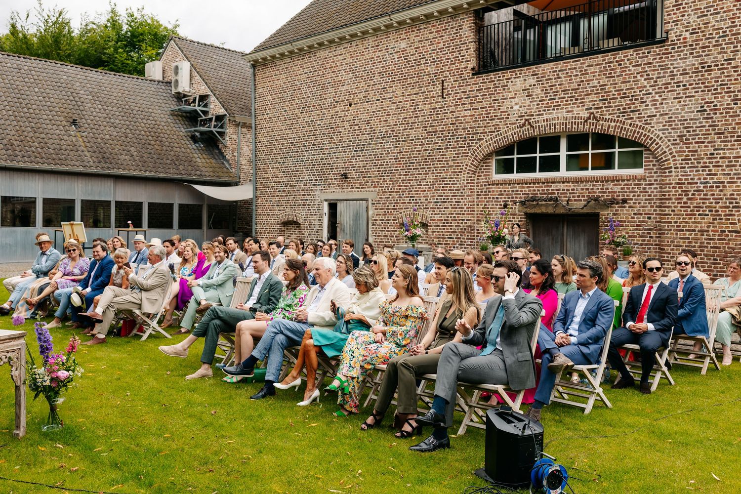 Wedding guests seated in white chairs on lawn during outdoor ceremony at rustic brick venue.