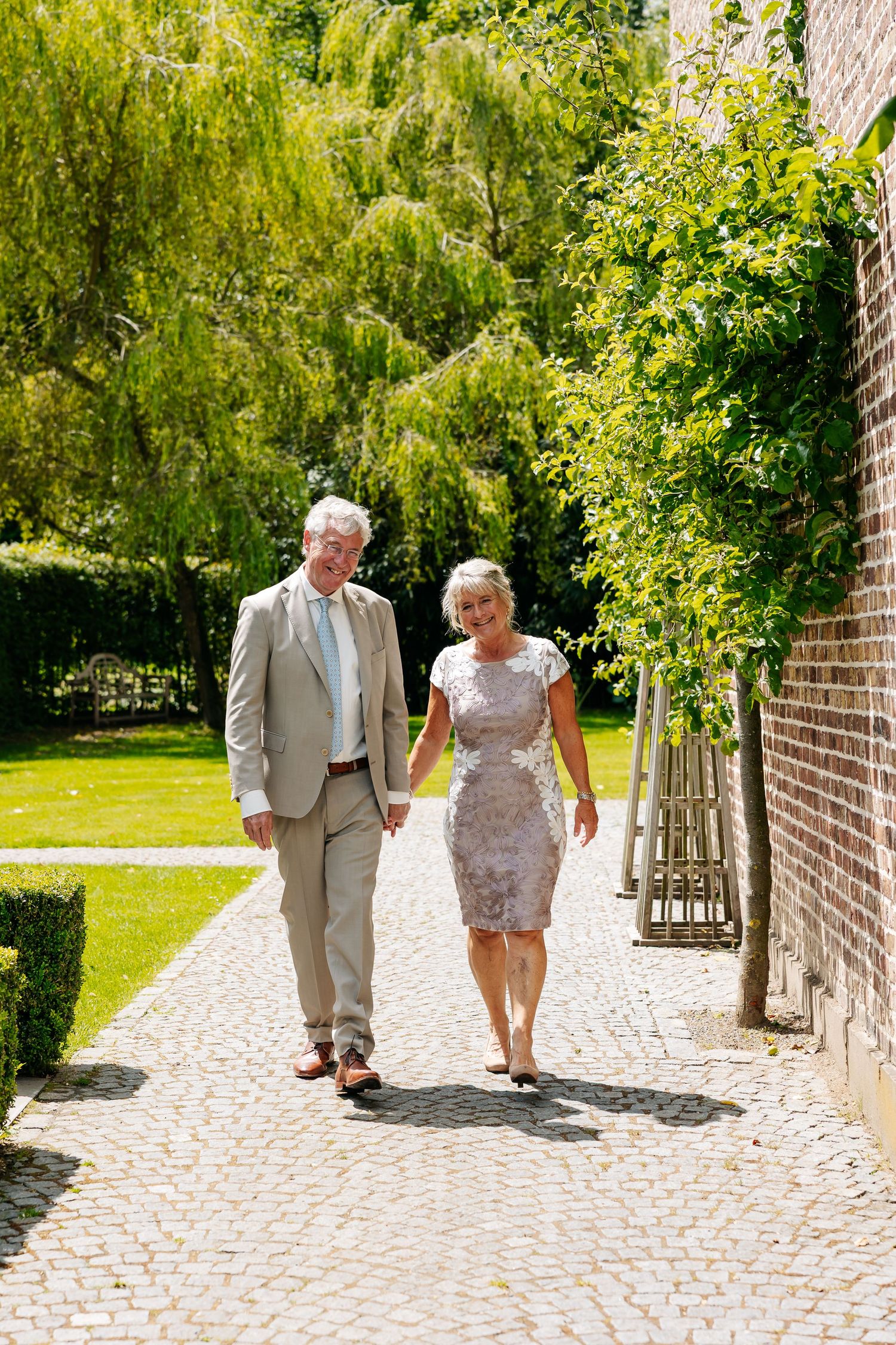 Elegant older couple walks hand in hand along a cobblestone path through a sunny garden setting.