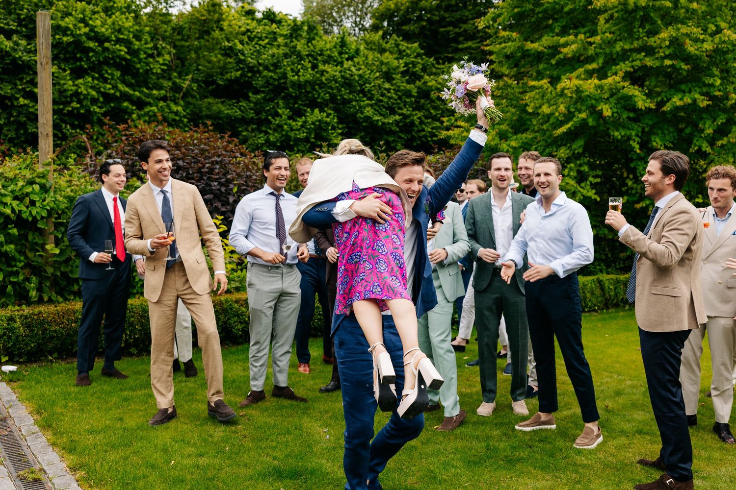 Excited wedding guests lift a woman in a purple dress during outdoor celebration on grass.