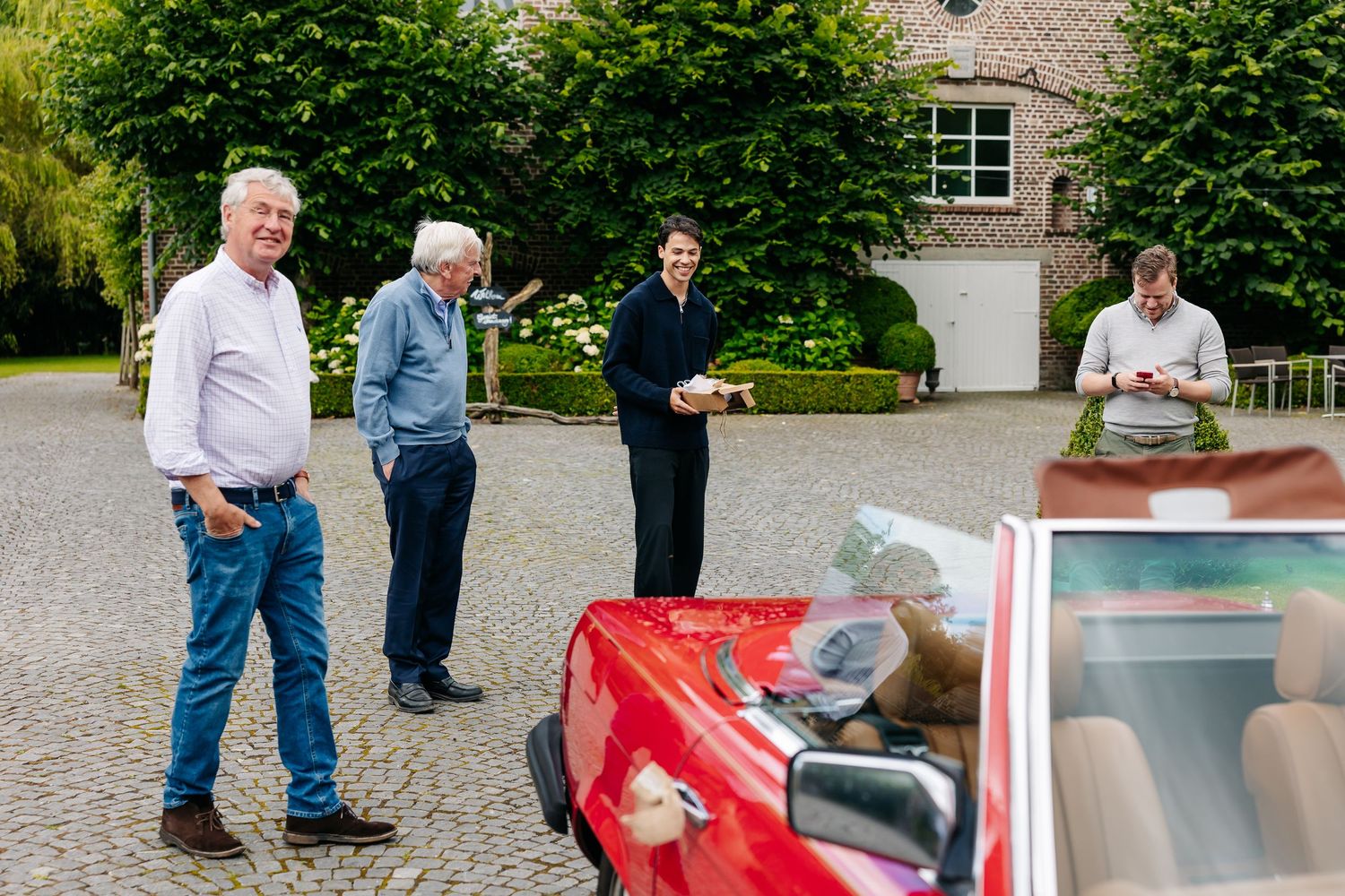 Group of men gathered around a classic red convertible car in a cobblestone courtyard.