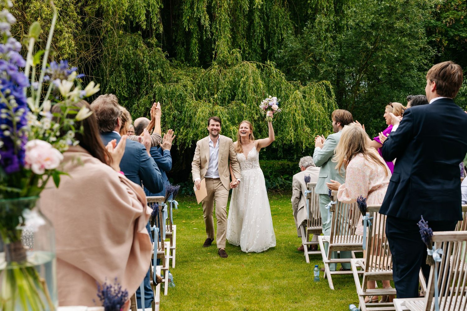 Joyful newlyweds walk down outdoor aisle raising arms as wedding guests applaud in lush garden.