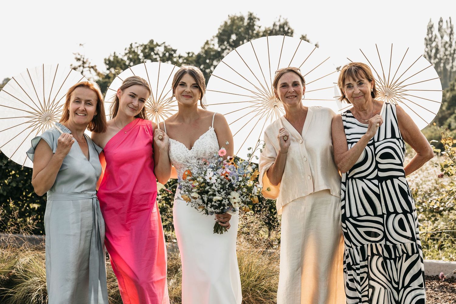 Bride and four bridesmaids holding white parasols posing together outdoors at a sunny wedding celebration.