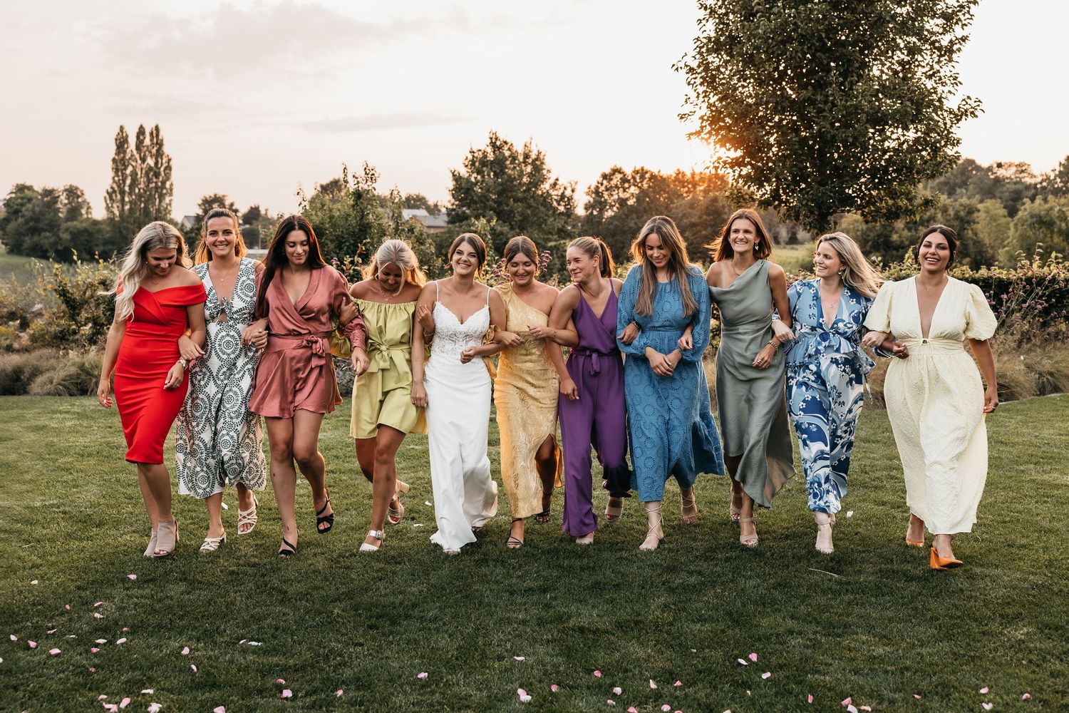 Joyful bride and female wedding guests running together in colorful dresses at sunset.