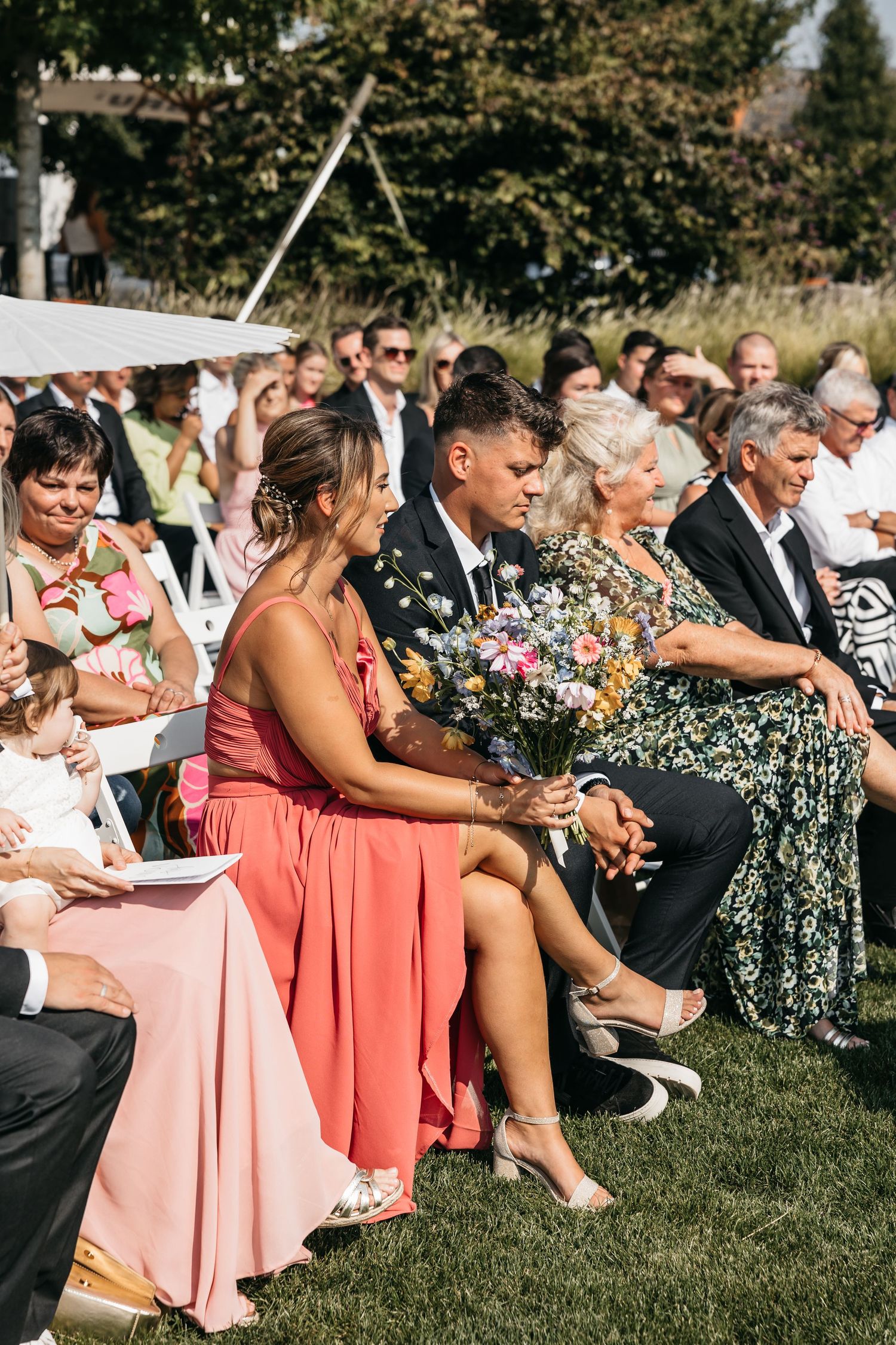 Wedding guests seated outdoors holding colorful wildflower bouquet during sunny ceremony.