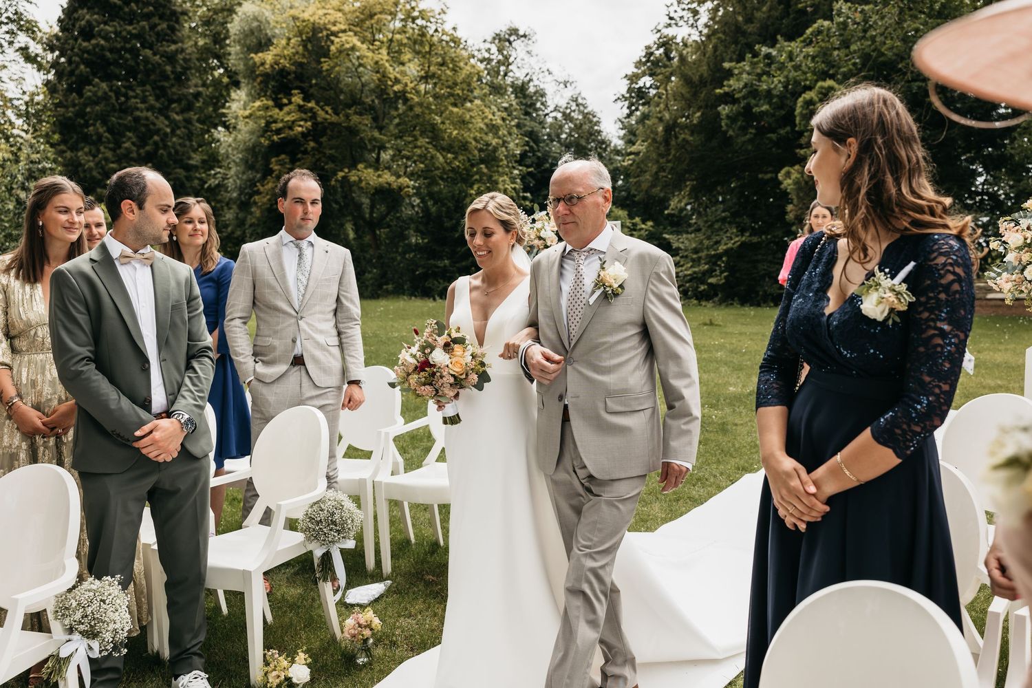 Bride walks down outdoor aisle with escort past smiling guests at sunny garden wedding ceremony.