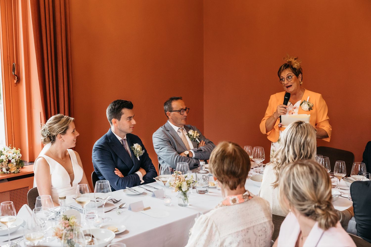 Woman in orange jacket delivers speech to bride and groom at wedding reception dinner.