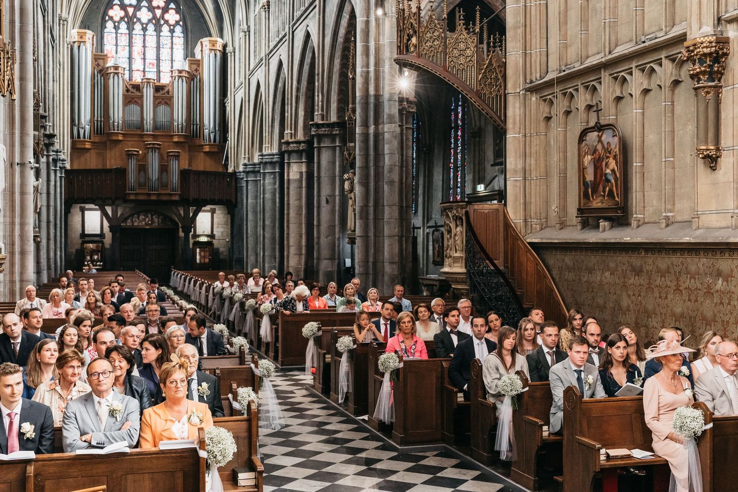 Large congregation of wedding guests seated in wooden pews of grand Gothic cathedral during ceremony.