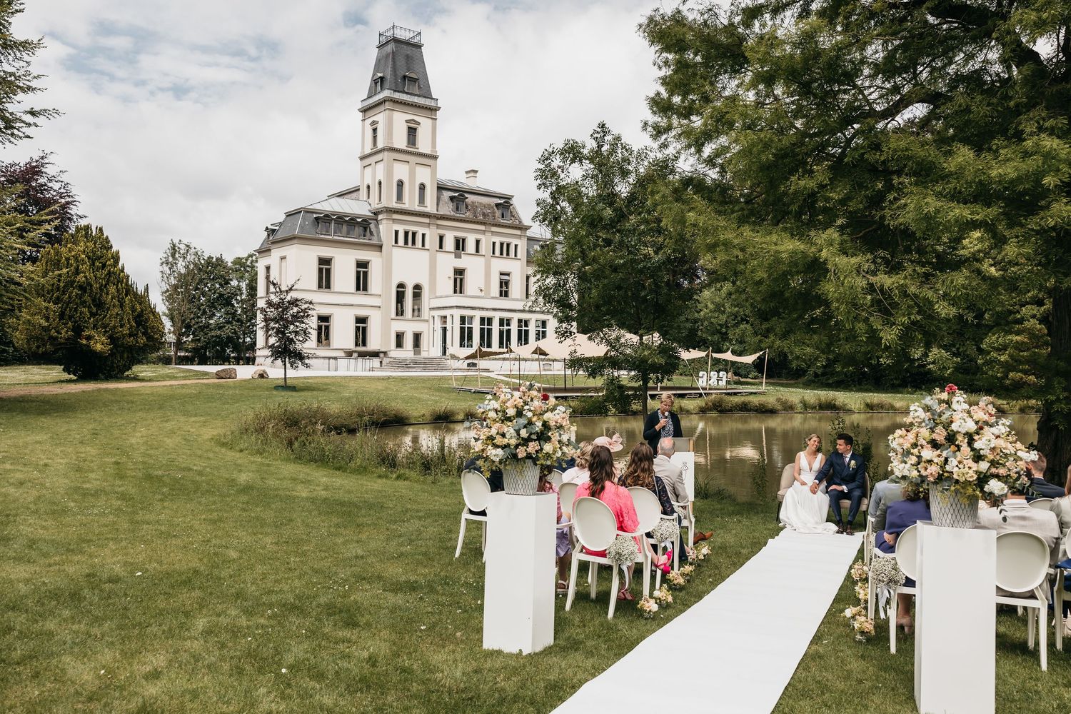 Outdoor wedding ceremony setup with white aisle and floral arrangements before grand estate.