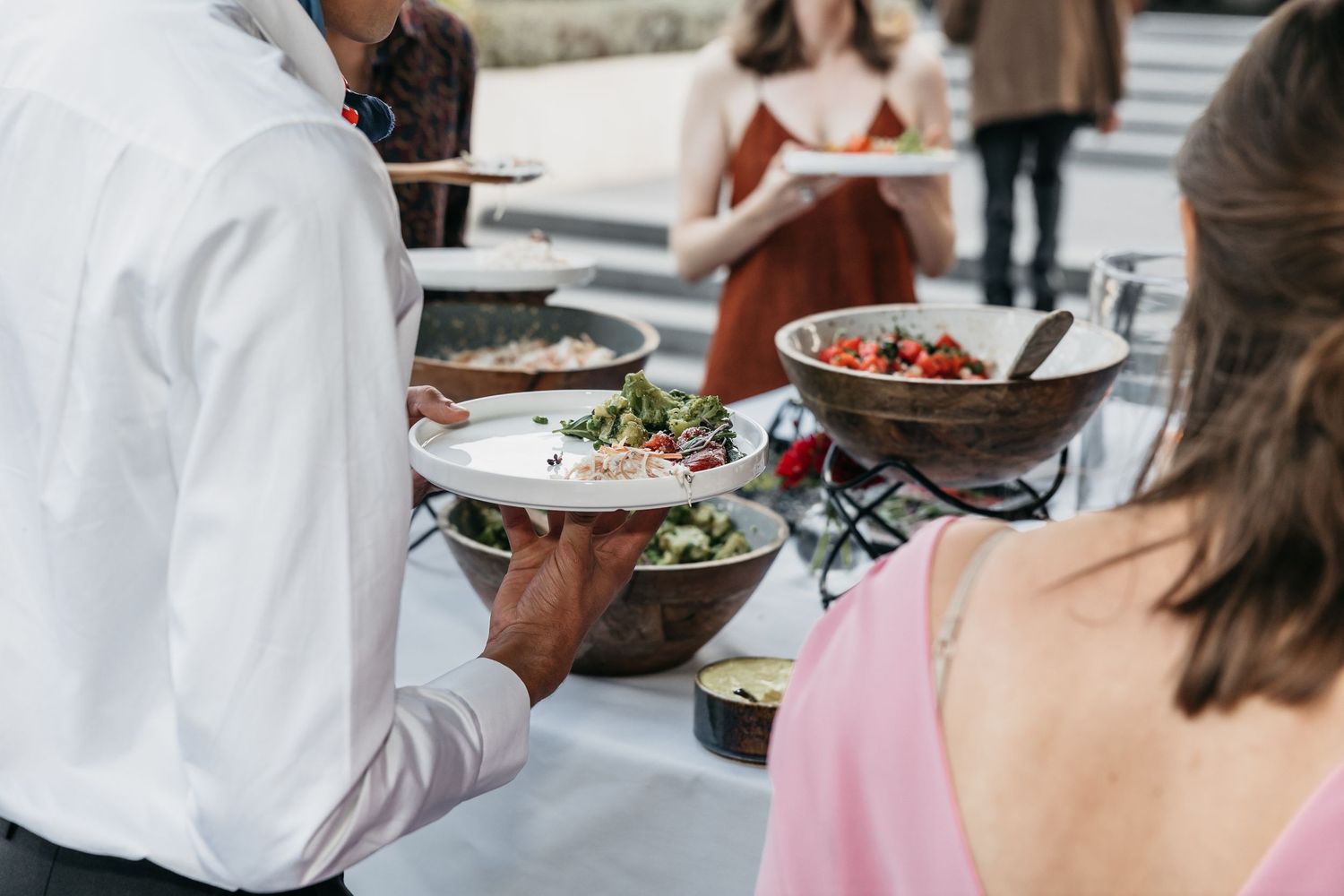 Guests serving themselves fresh salads from large wooden bowls at an elegant outdoor catered event.