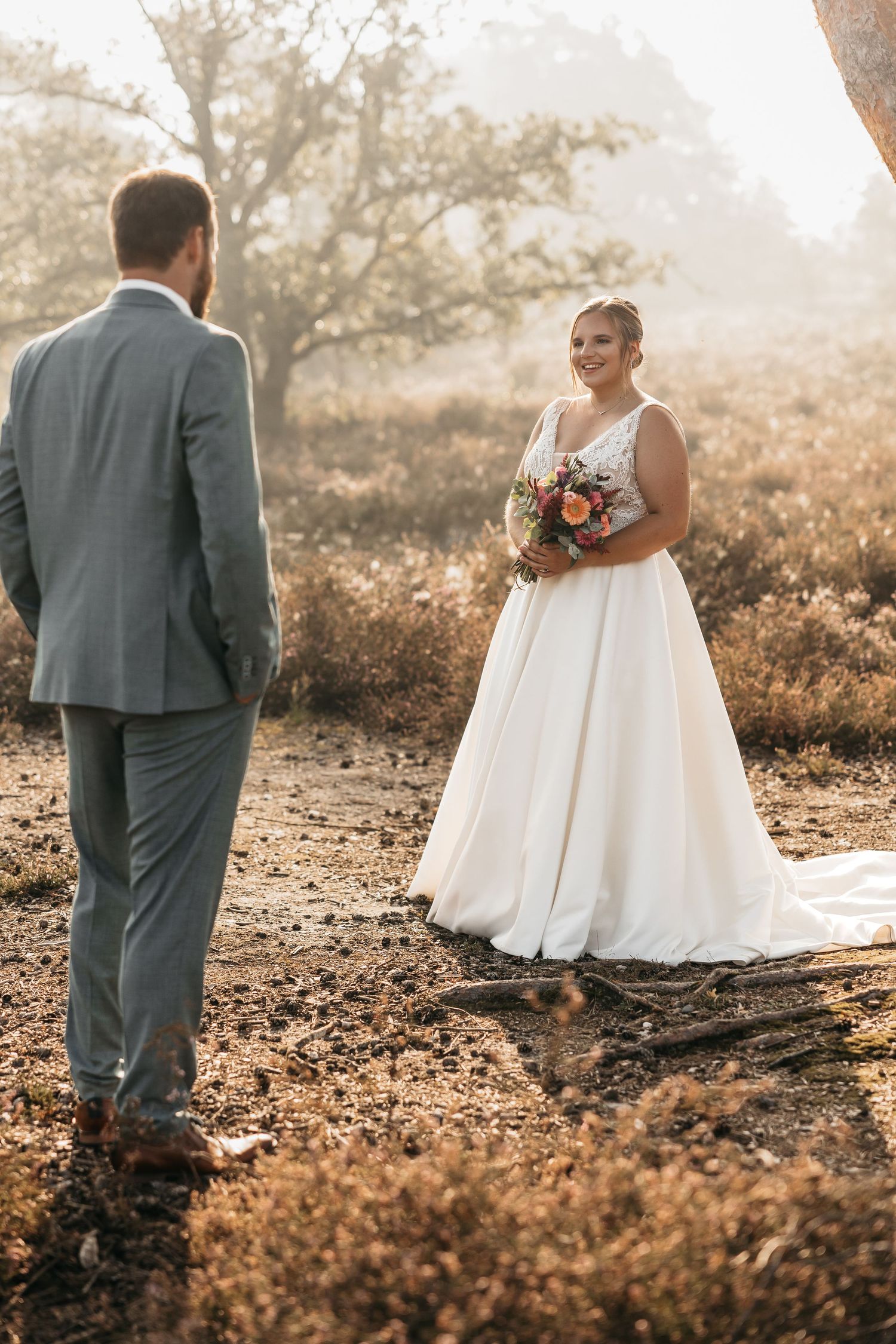 Smiling bride in white gown holds colorful bouquet while facing her groom in a sun-drenched autumn heathland.