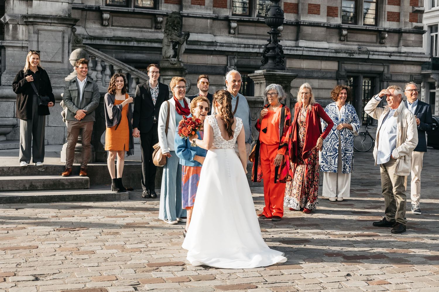 Bride and groom posing with wedding guests in colorful attire on sunny plaza steps.