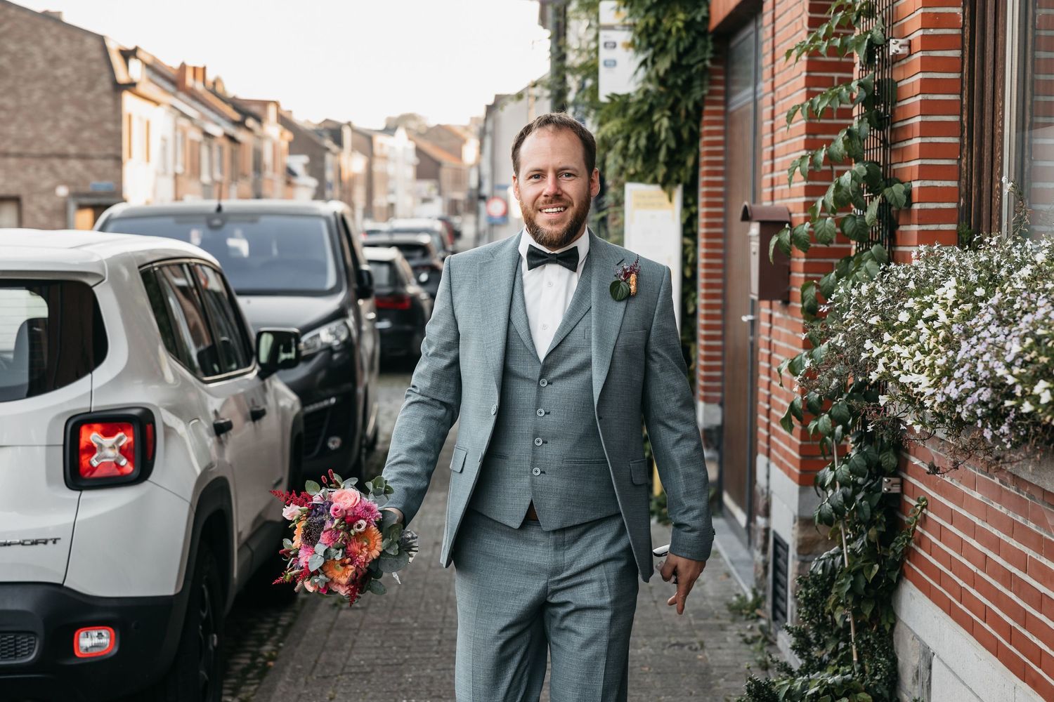 Smiling groom in grey three-piece suit holding a colorful bridal bouquet on a street.