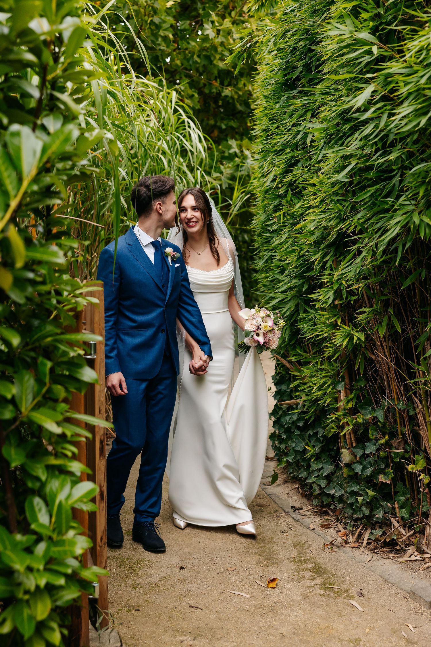 Bride and groom walking hand in hand through a lush green bamboo garden path on wedding day.
