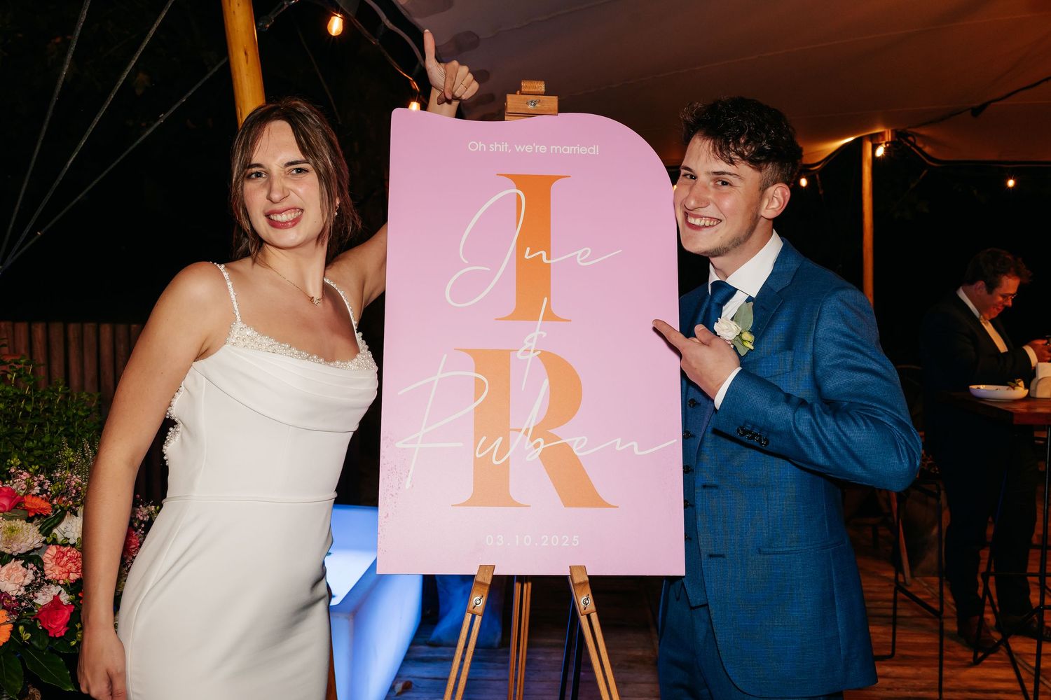 Smiling bride and groom standing next to a pink and gold monogram wedding welcome sign.