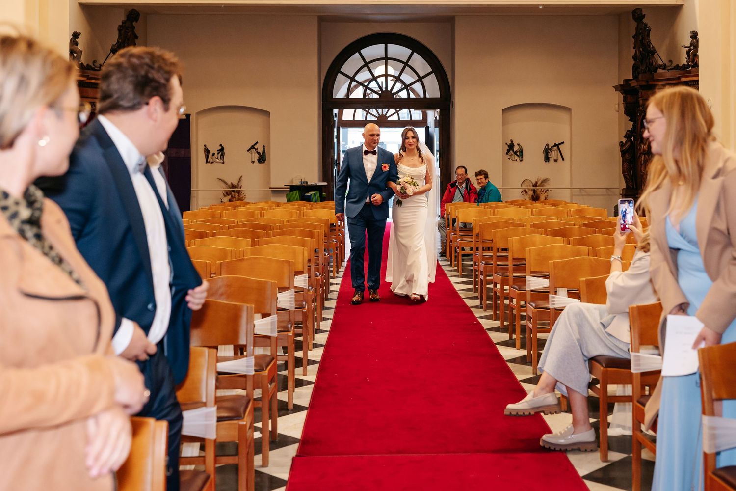 Bride in white gown being escorted down red carpet aisle by father in church wedding ceremony.