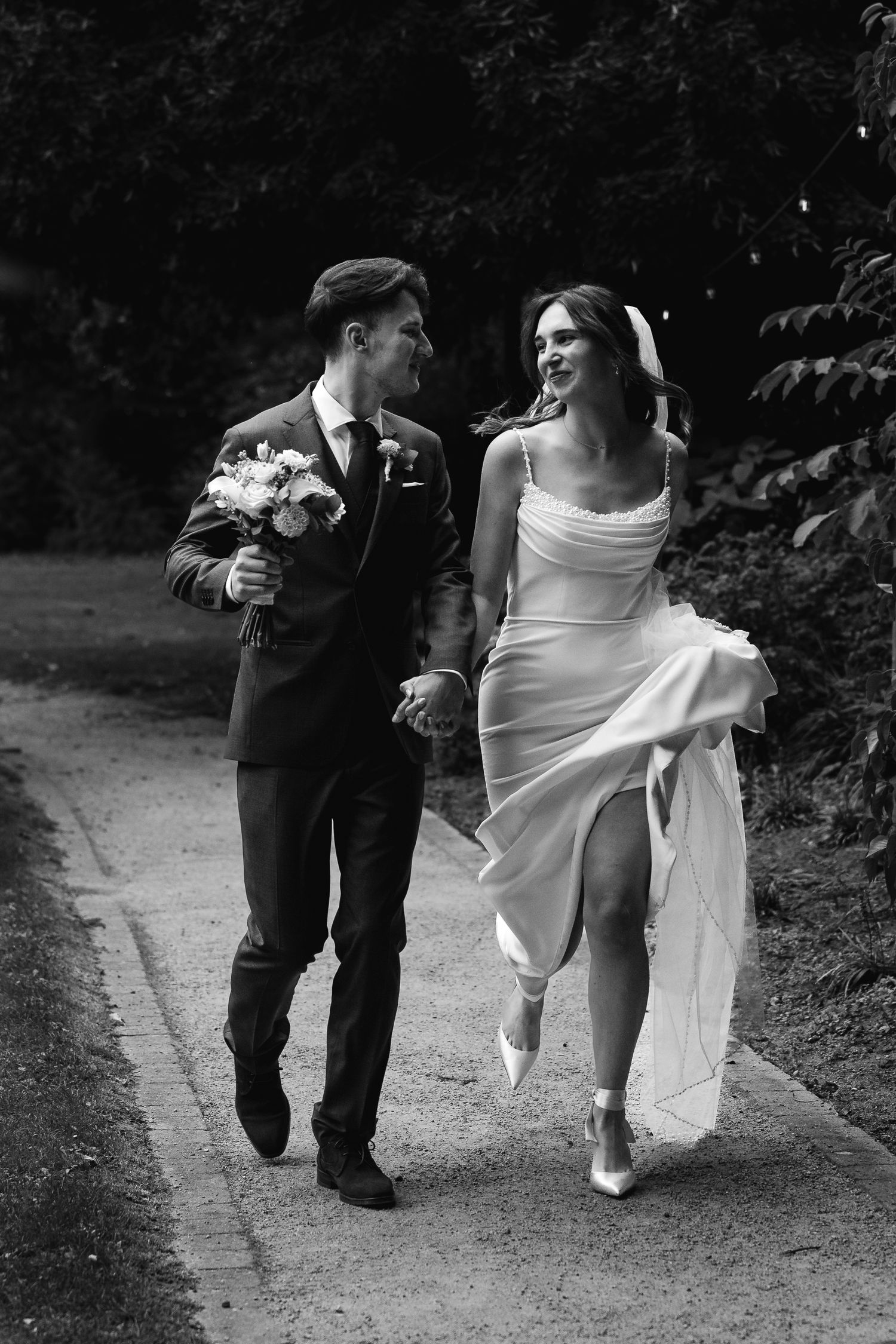 Black and white photo of smiling bride and groom walking hand in hand through a garden path.