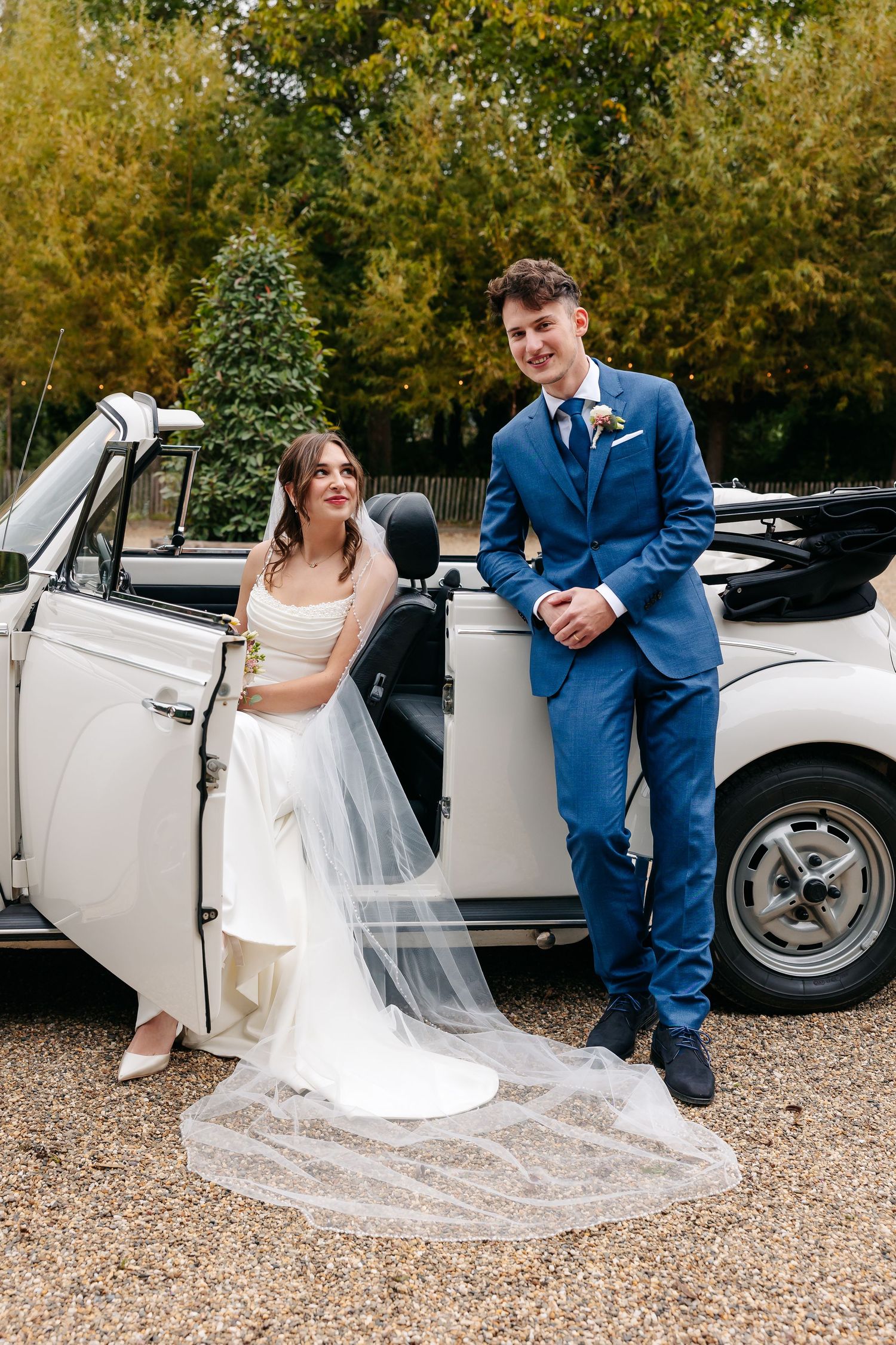 Bride in white gown with long veil and groom in blue suit posing beside a vintage white convertible car.