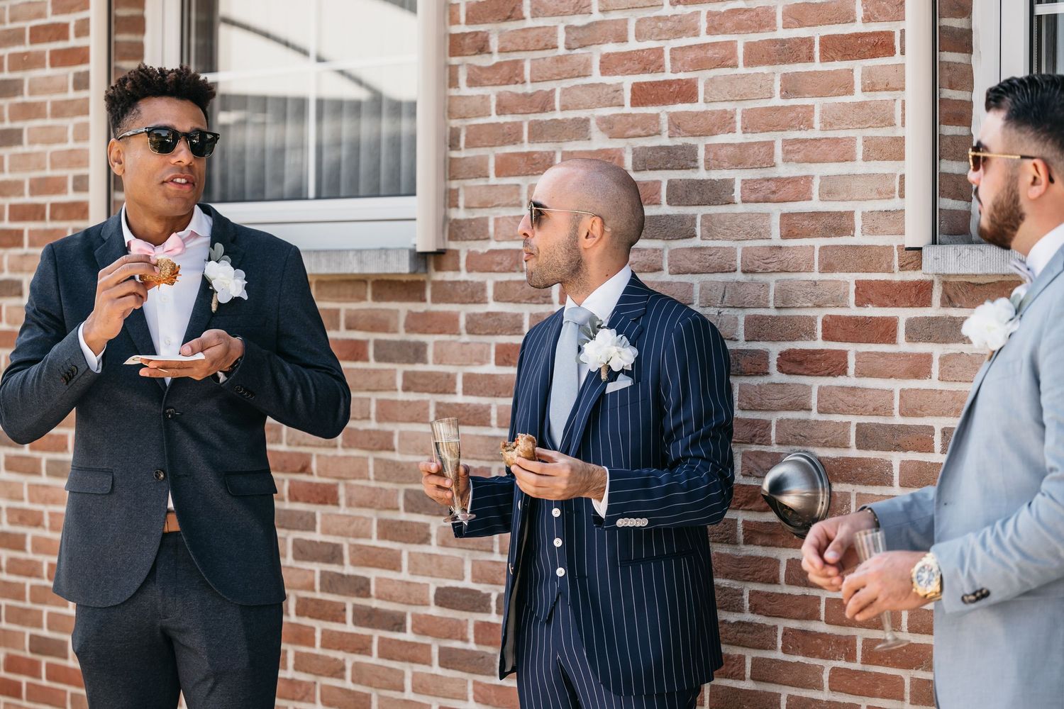 Three groomsmen in suits with boutonnieres socializing outside a brick building at a wedding reception.