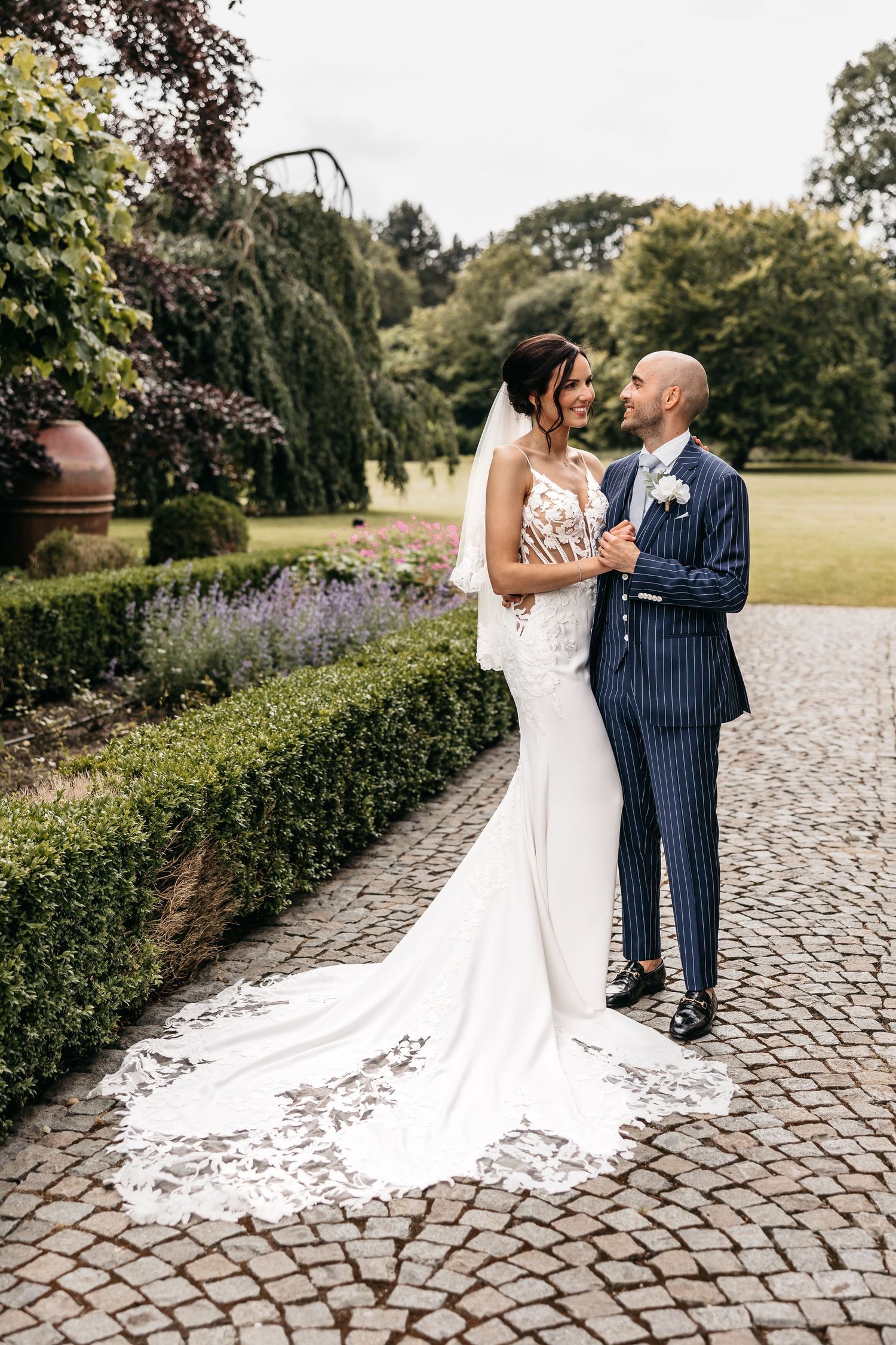 Bride in elegant lace train gown and groom in pinstripe suit pose on a cobblestone garden path.