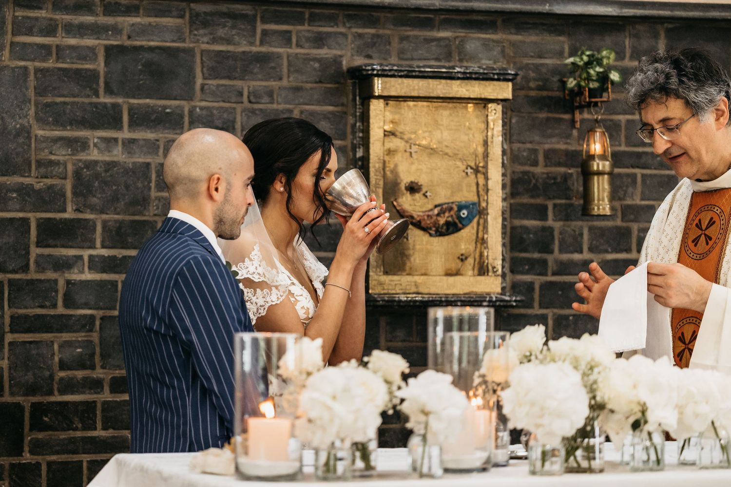 Bride drinking from ceremonial chalice as groom and priest look on during Catholic wedding mass.