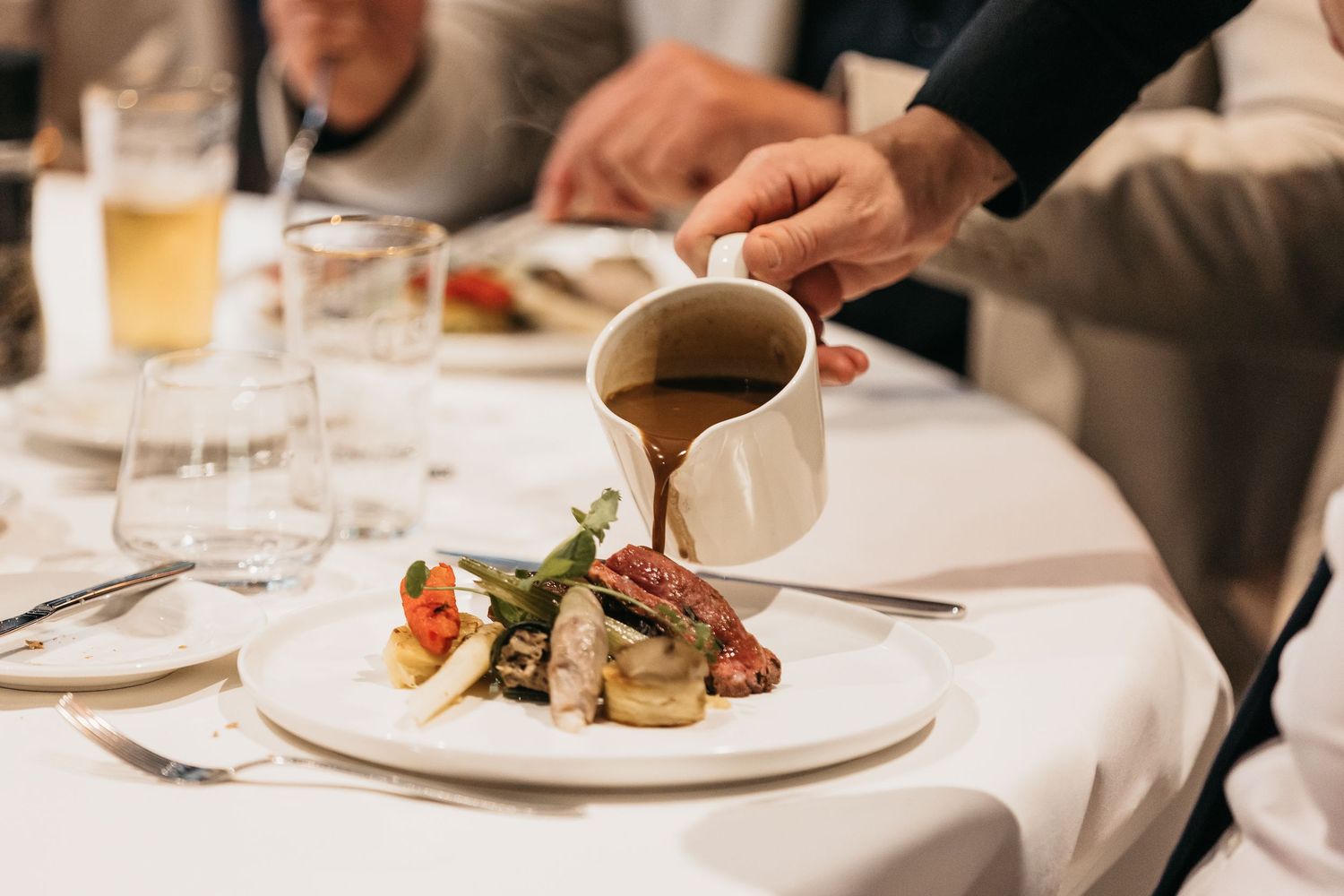 Waiter pouring rich brown gravy sauce over elegant wedding reception main course on white plate.