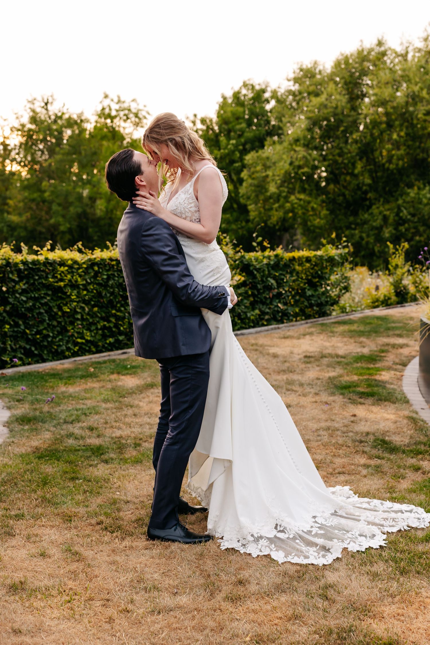 Groom lifts bride in a white lace wedding gown as they share a tender moment in a garden at sunset.