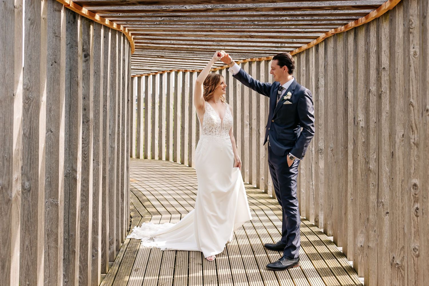 Groom twirls his bride in a white gown through a rustic wooden walkway on their wedding day.