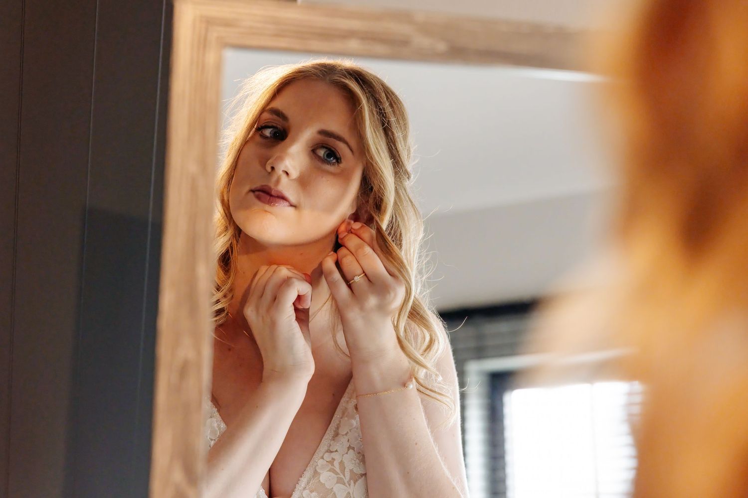 Blonde bride in white wedding dress puts on earrings while looking in a mirror during bridal preparations.