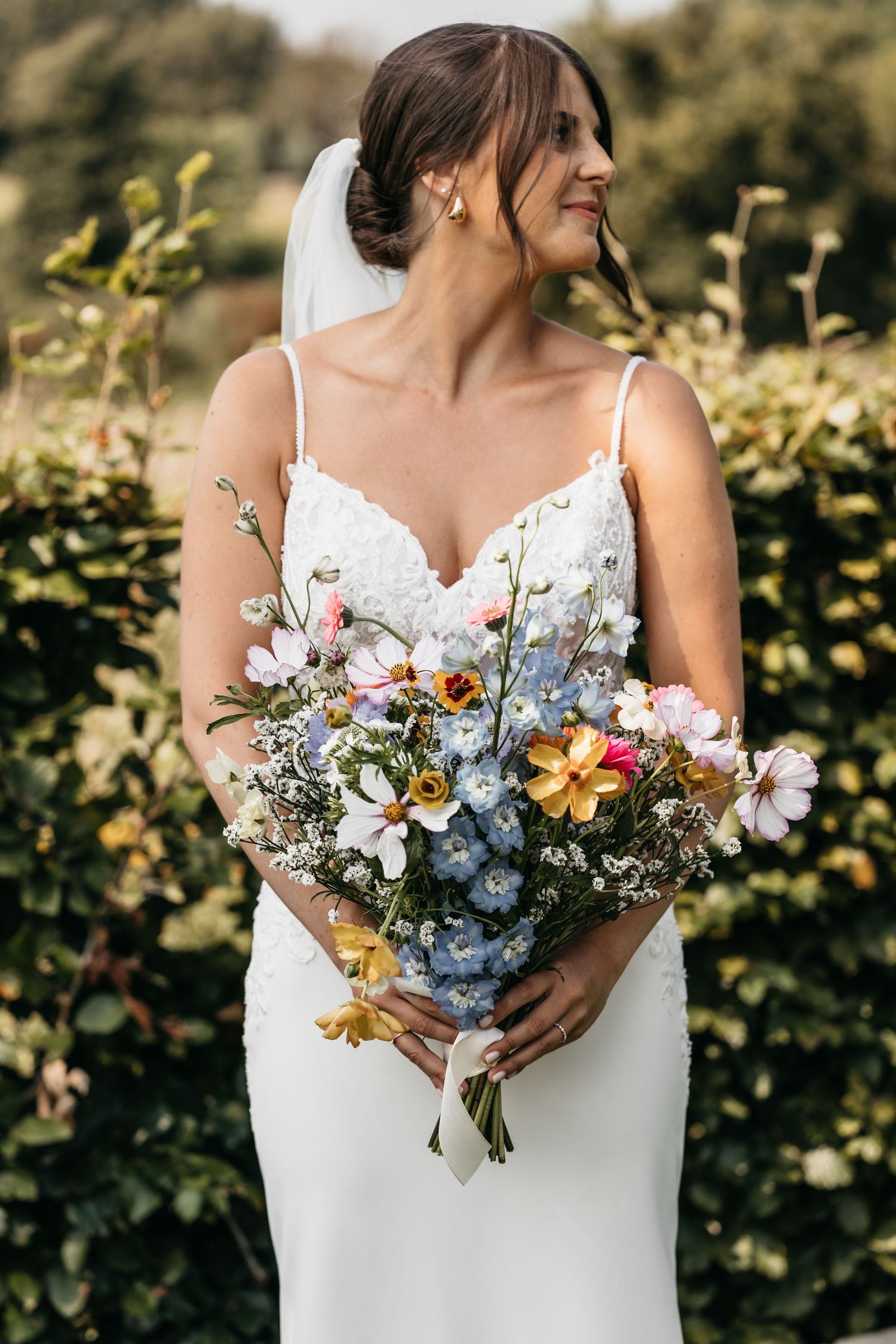Bride in spaghetti-strap lace gown holding colorful wildflower bouquet with veil.