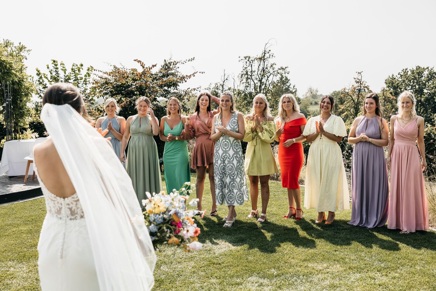 Bride in white gown and veil faces colorful bridesmaids in mismatched dresses during outdoor wedding bouquet toss.
