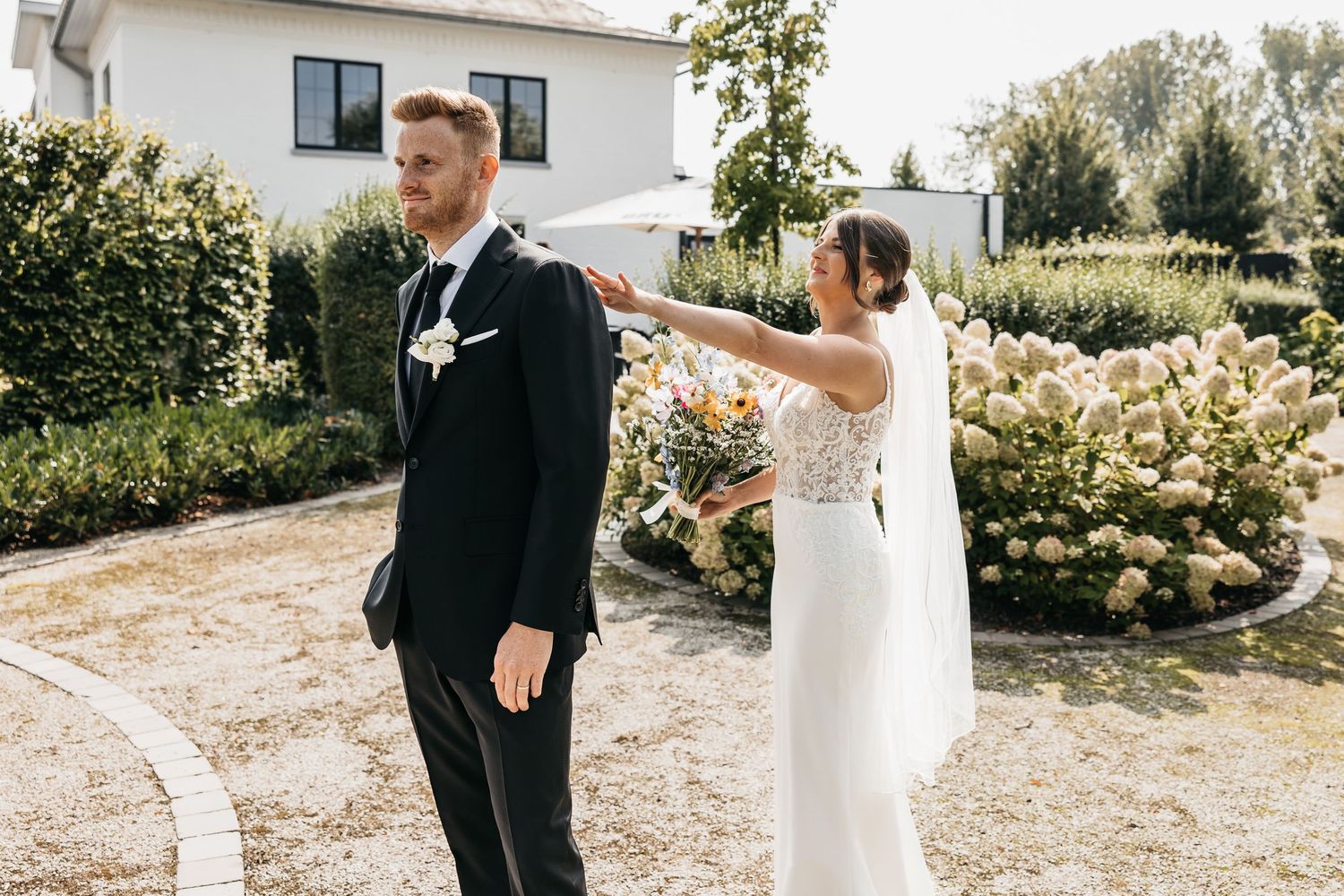 Bride taps groom on shoulder during first look moment in sunny garden with hydrangeas.