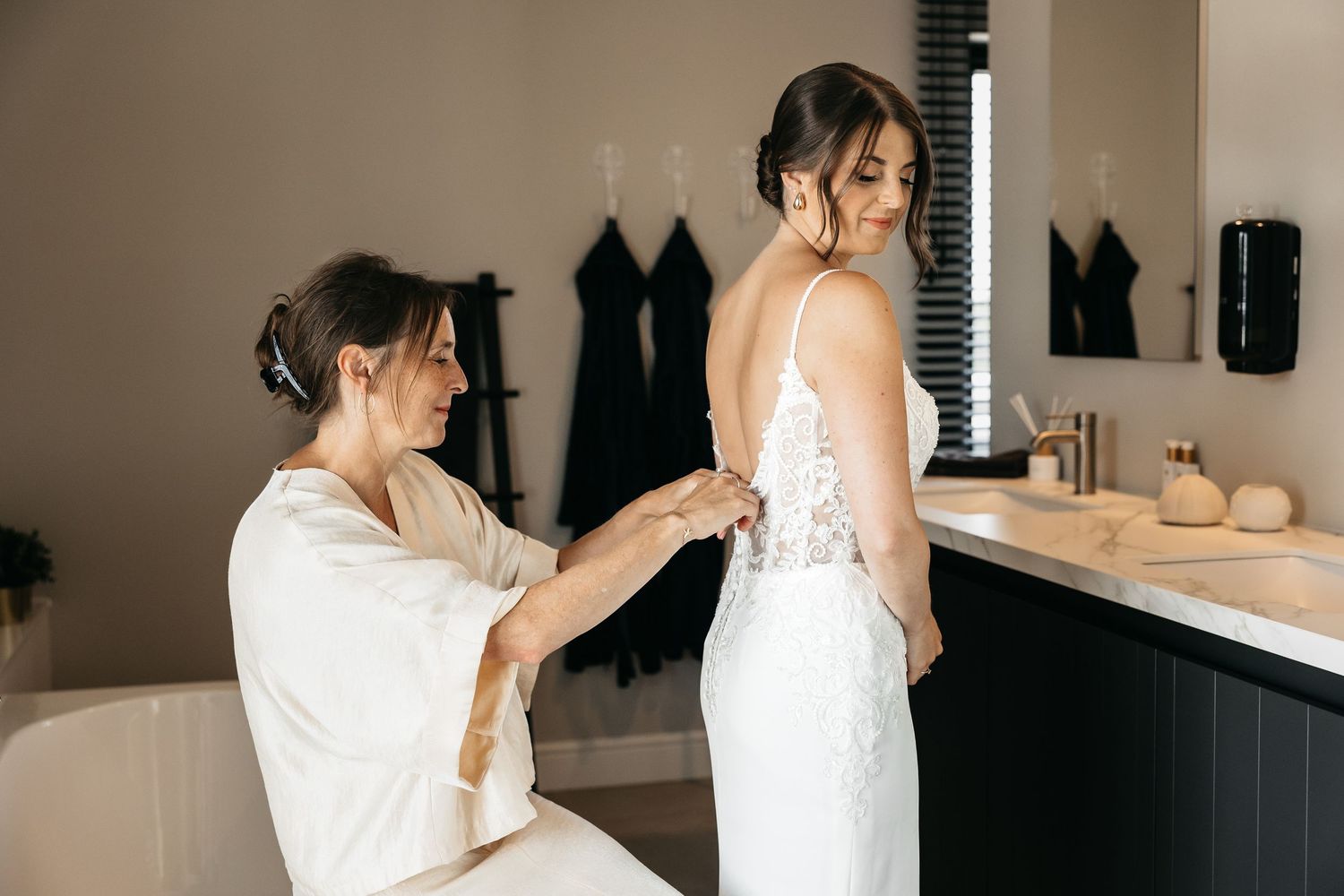 Woman in white robe helping bride fasten the back of her lace wedding dress.