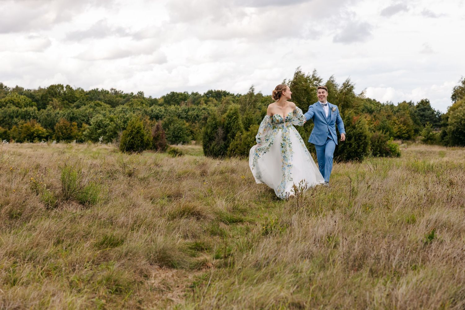 Bride and groom walking hand in hand through a wide open grassy field under a cloudy sky.