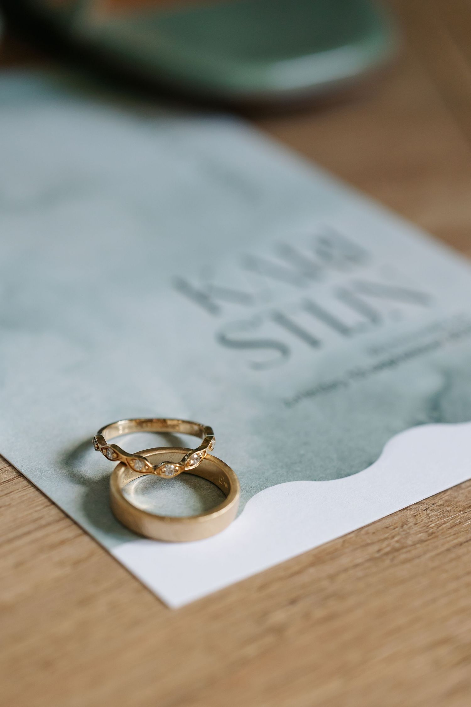 Two gold wedding bands resting on watercolor wedding invitation card on wooden surface.